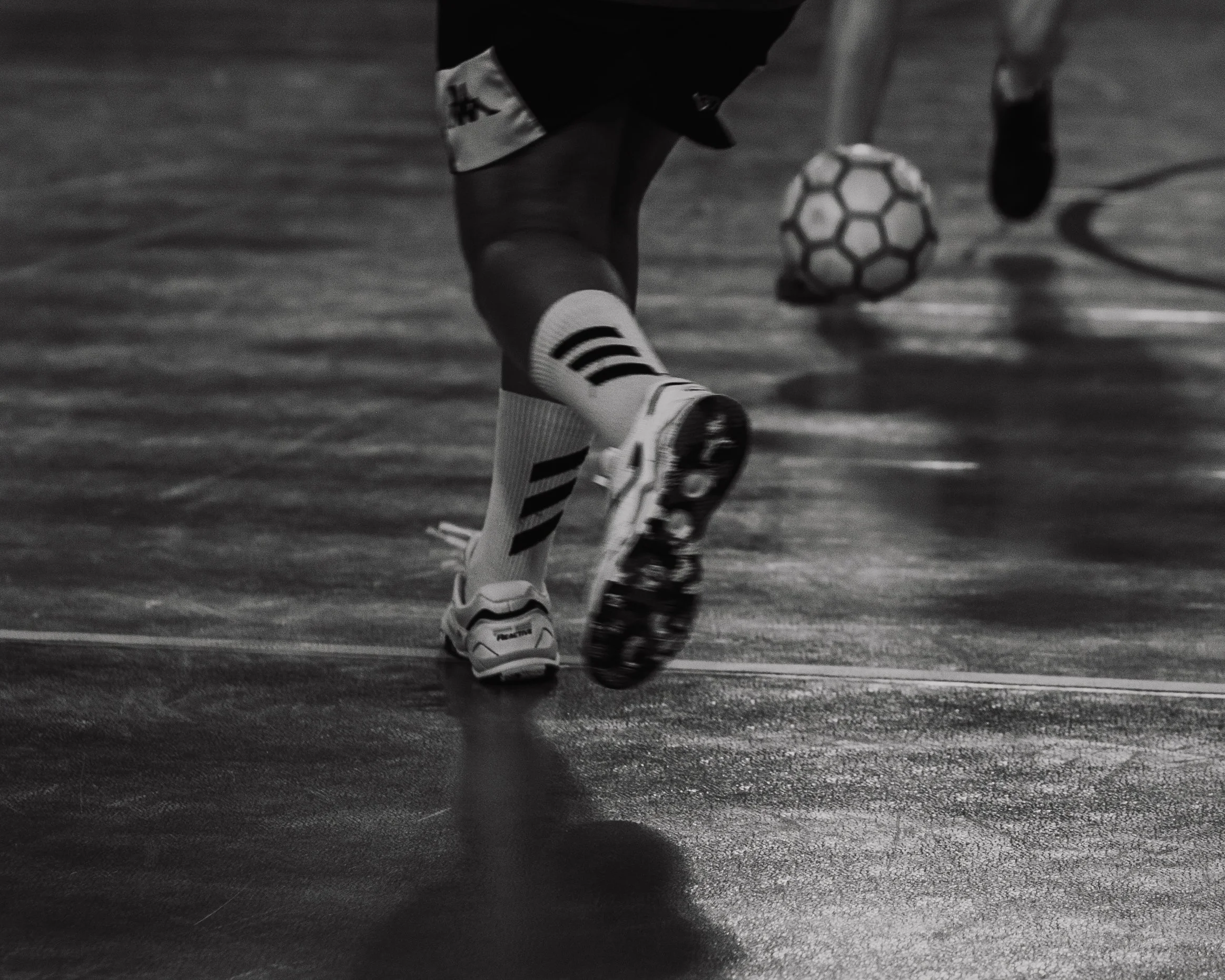 Jambes d'un joueur de football en train de courir sur un terrain sportif, avec un ballon de football sous un autre joueur en arrière-plan.