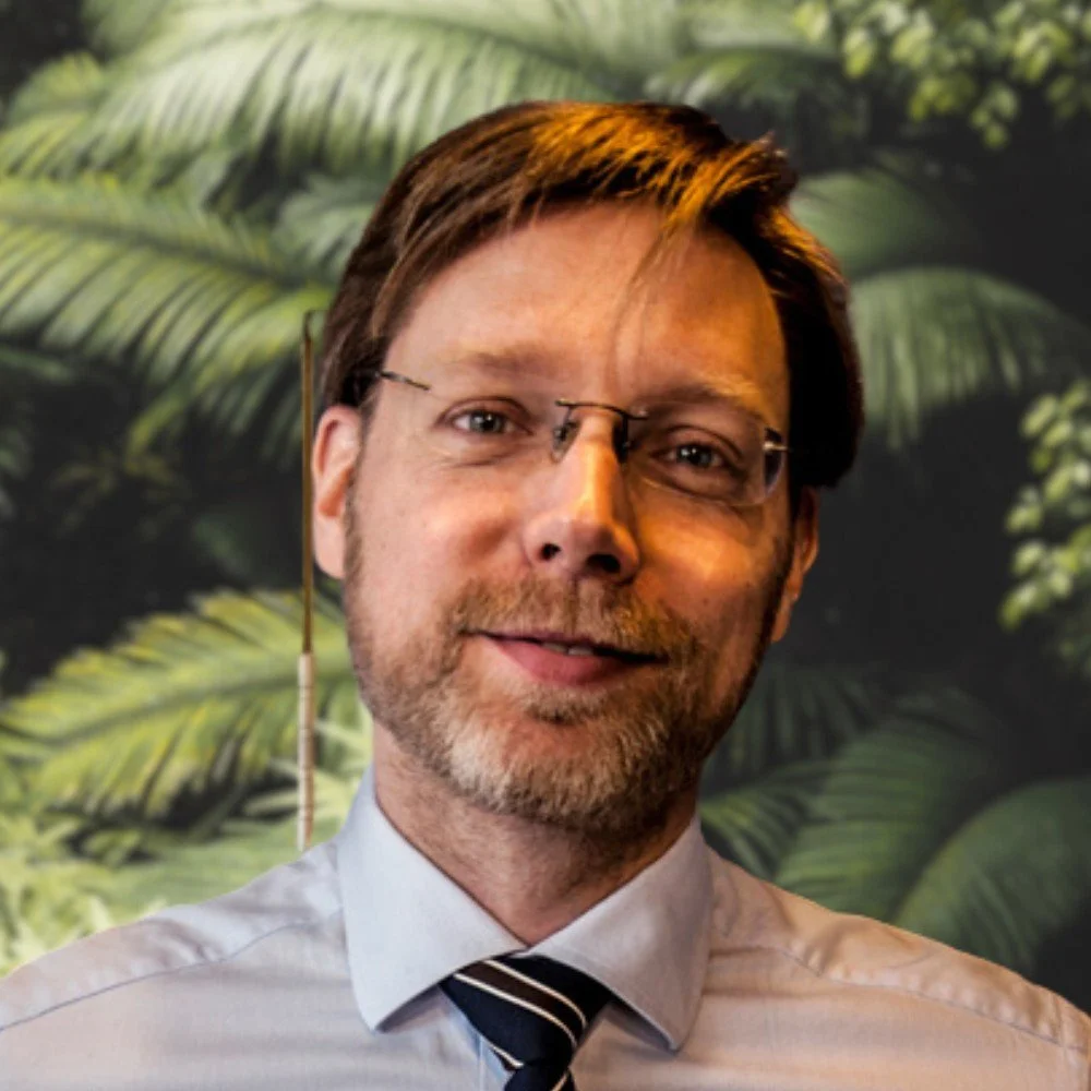 Man with glasses and beard, wearing a shirt and tie, with a leafy green background.