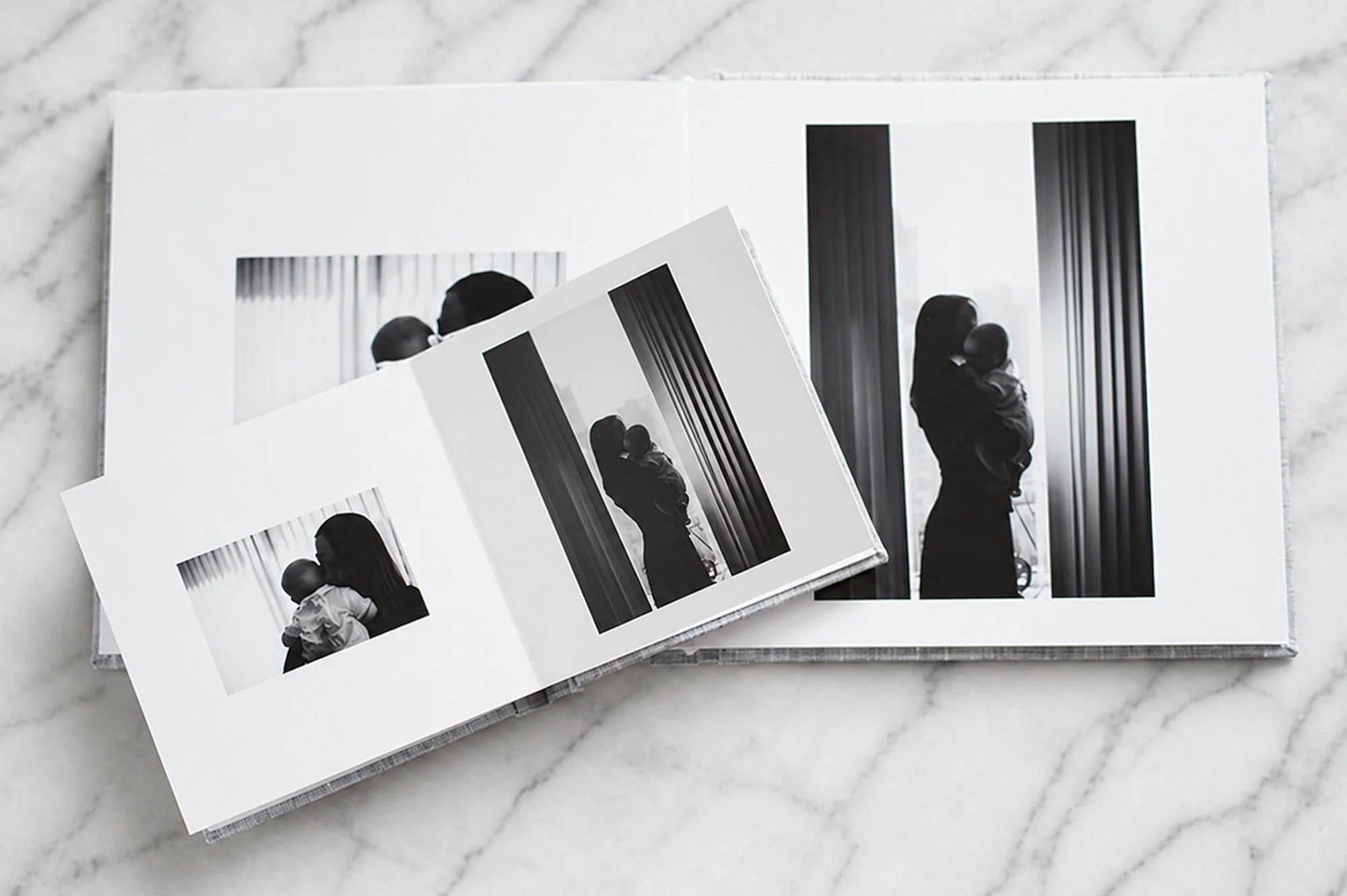 Fine art newborn albums in two sizes, open to black and white newborn photographs, linen covers, shown on marble surface.