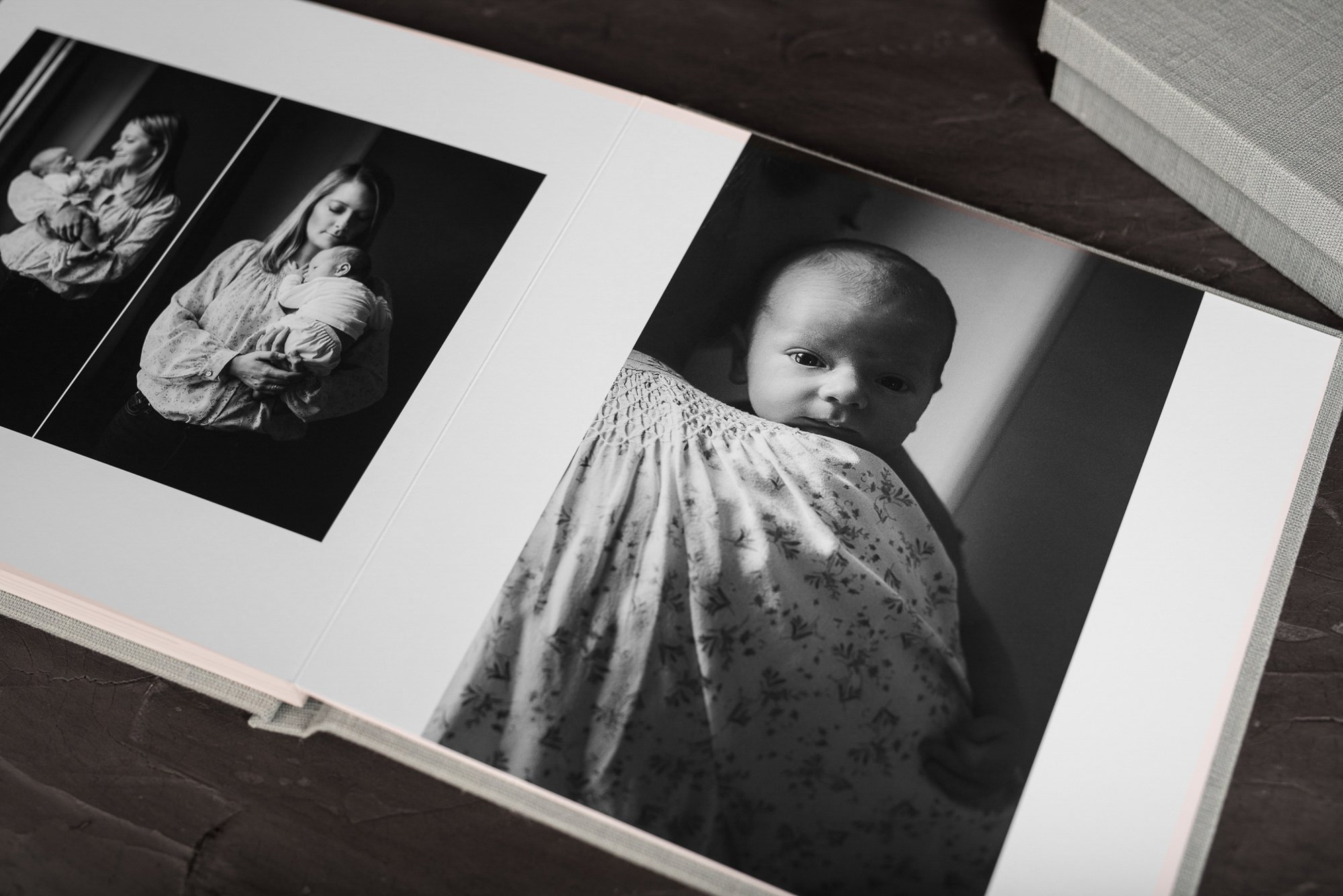Fine art newborn album open to black and white interior spread showing mother holding newborn and close portrait of infant, with matching linen keepsake box.