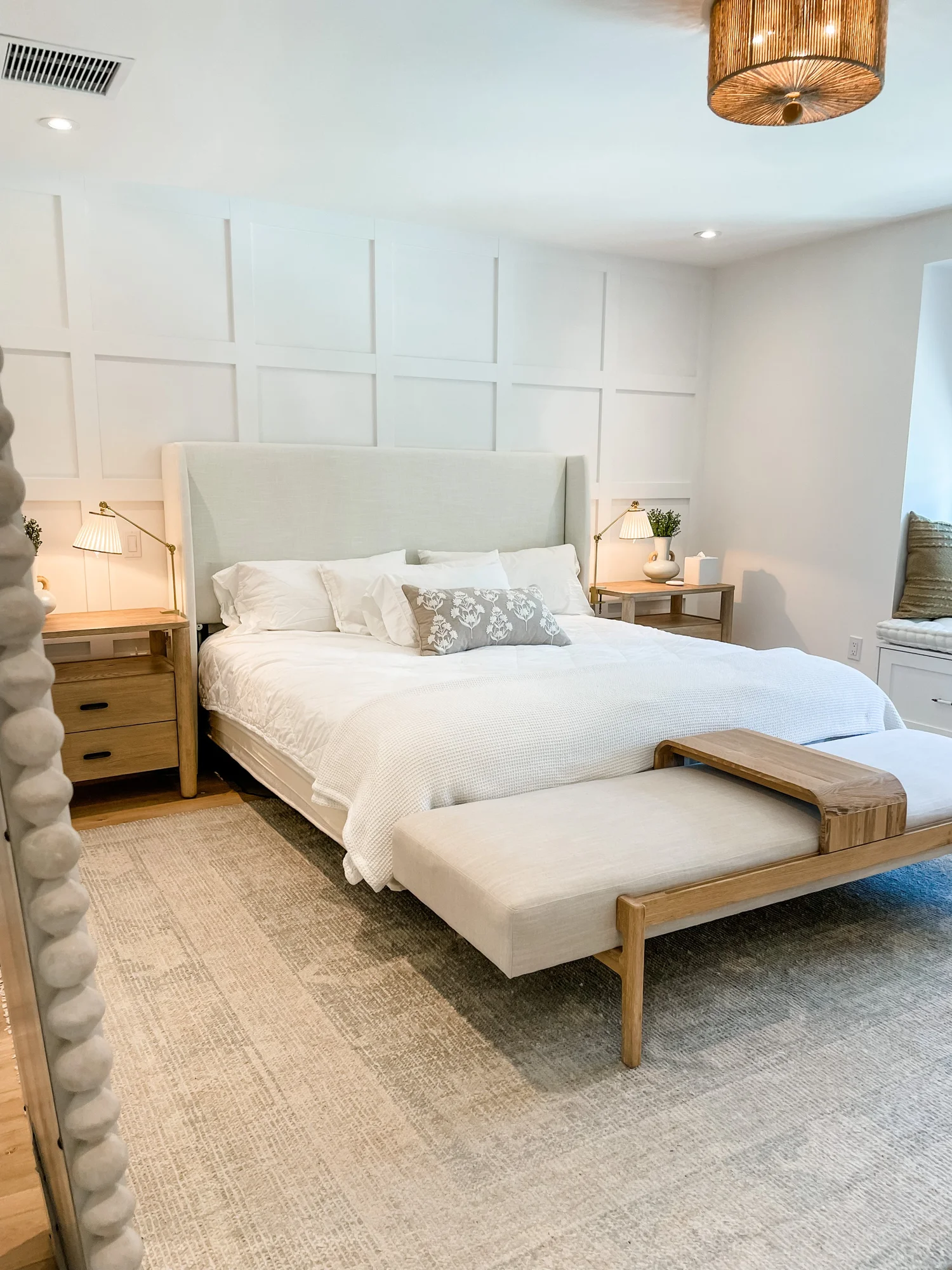 A calm bedroom with a paneled wall, soft bedding and a woven bench