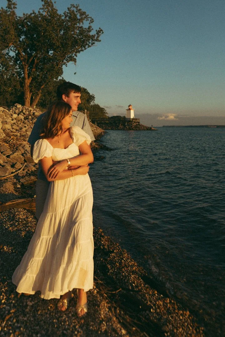 A young couple standing on a rocky shoreline during sunset, with a lighthouse in the background.