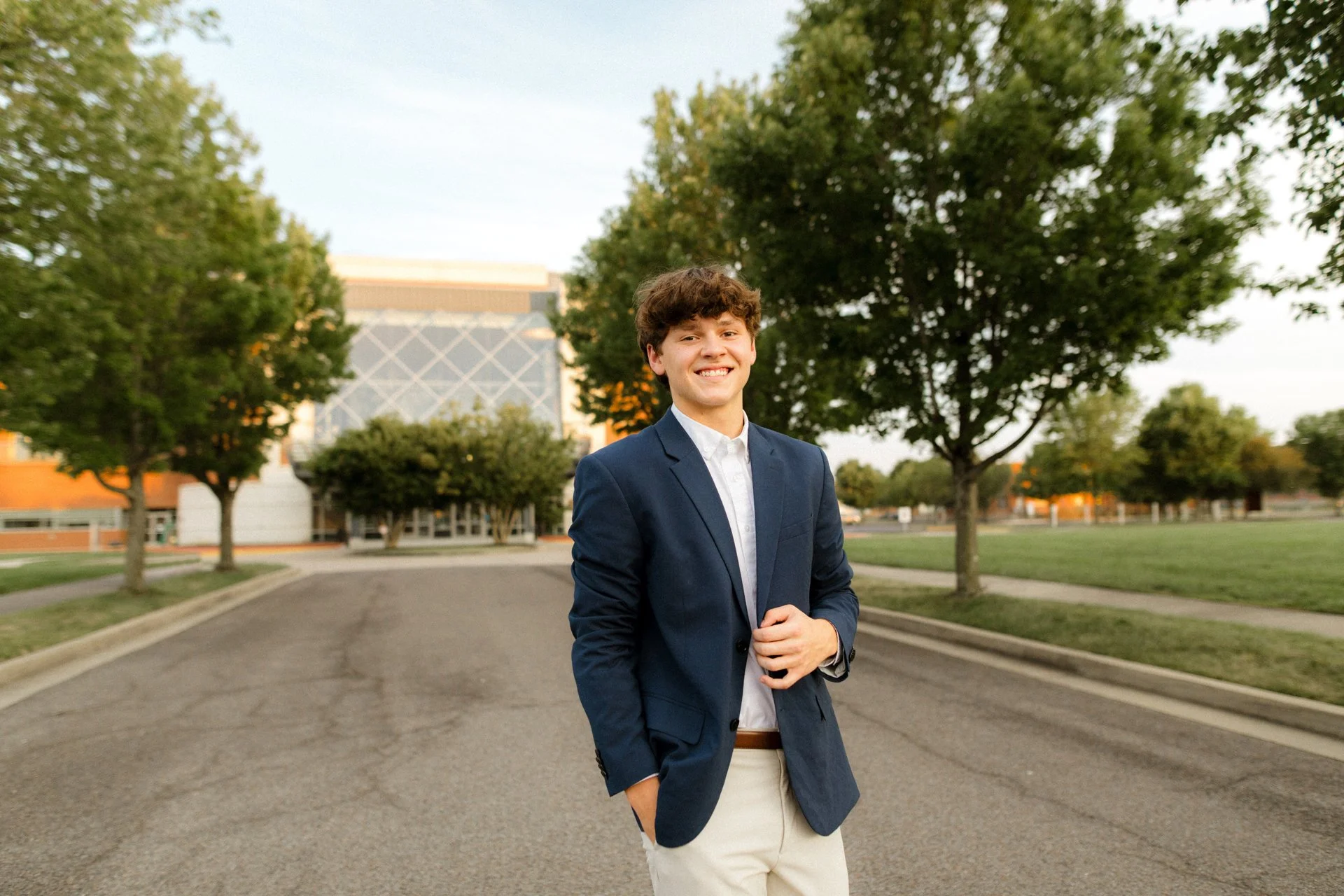 A young man in a navy blazer, white shirt, and beige pants standing on a paved road outdoors with green trees and a modern building in the background, smiling at the camera.
