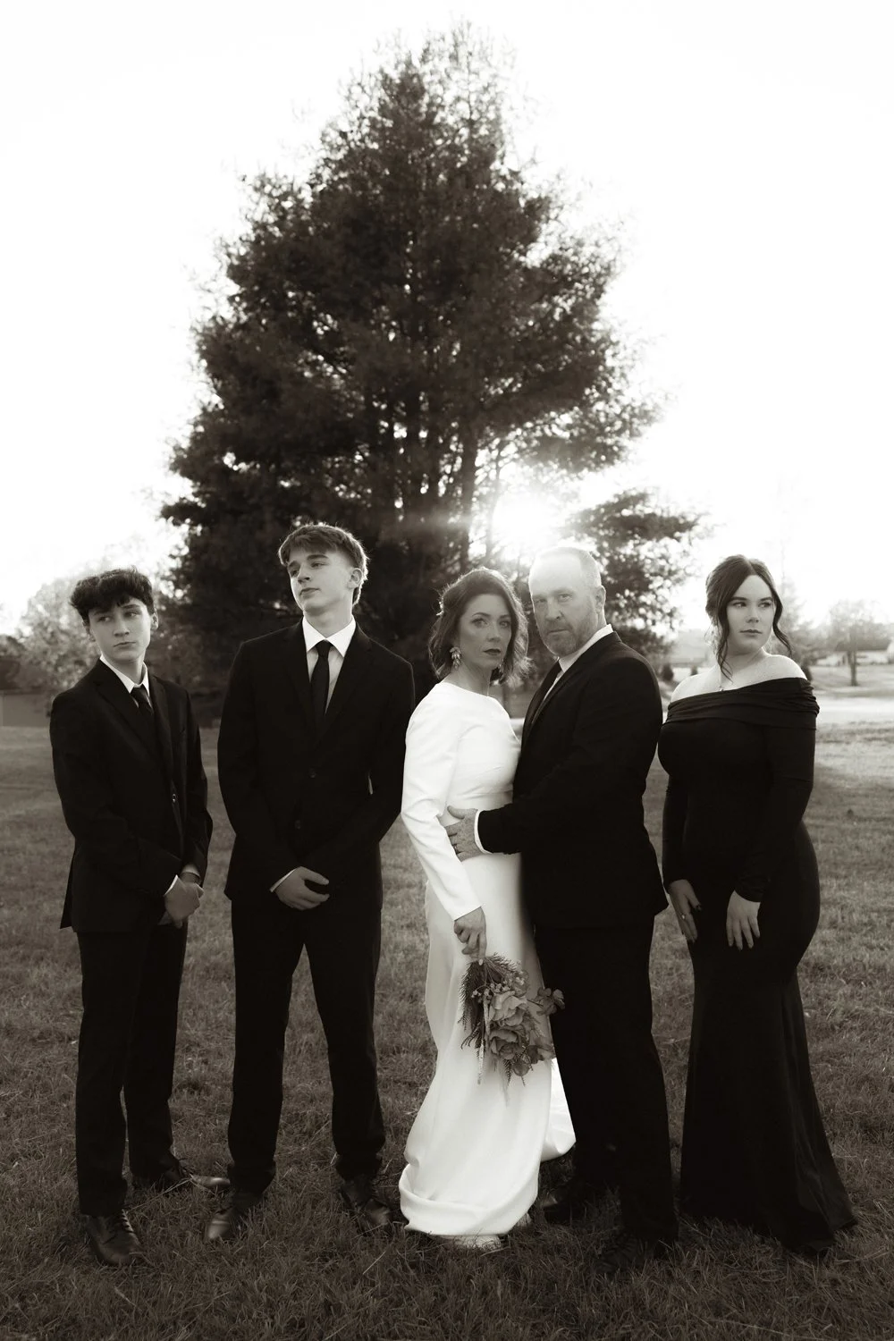 A black-and-white photo of a family dressed in formal attire outdoors near a large tree, with the sun setting behind them.