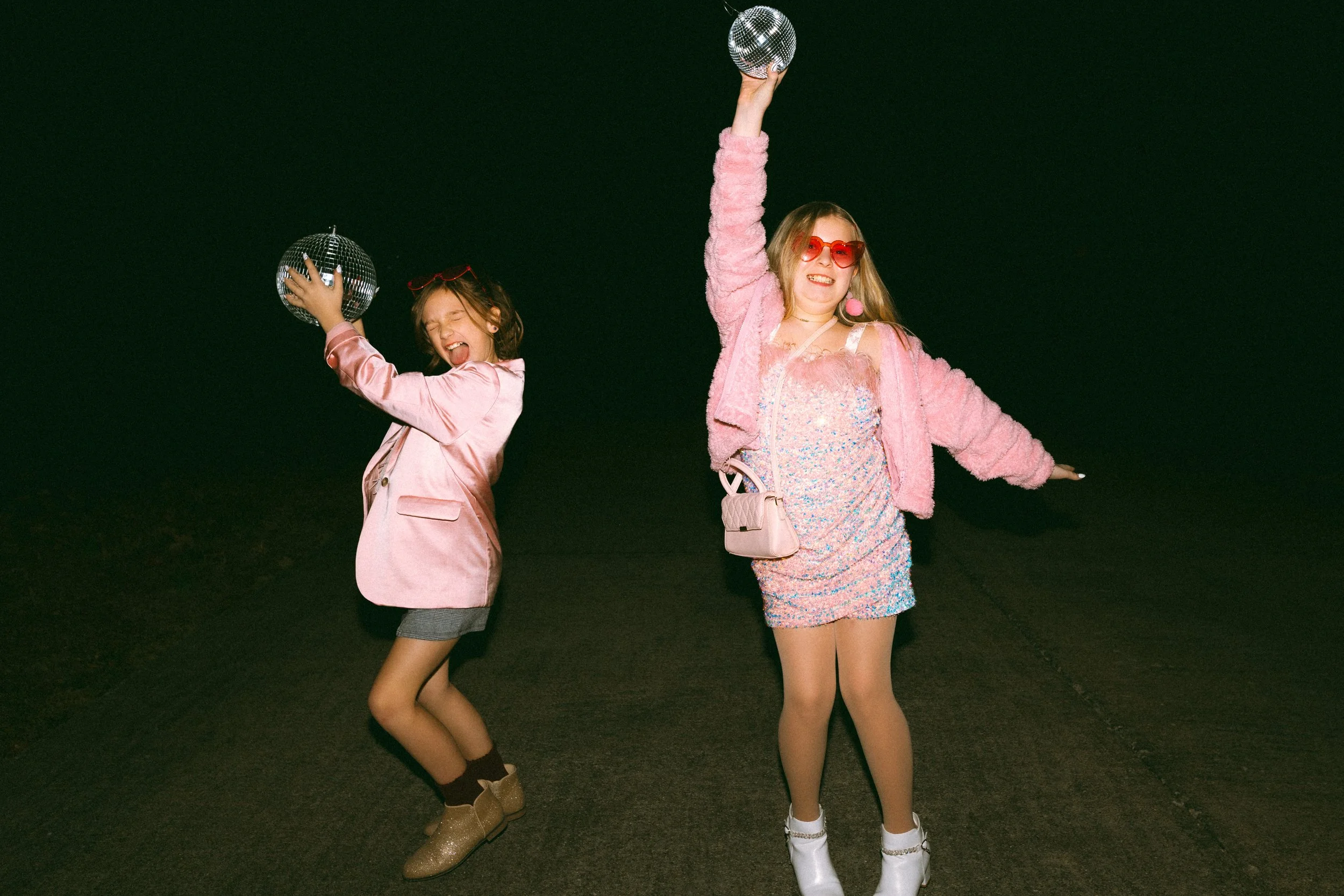 Two young women dressed in colorful, shiny outfits celebrating at night with disco balls, smiling and dancing outdoors.