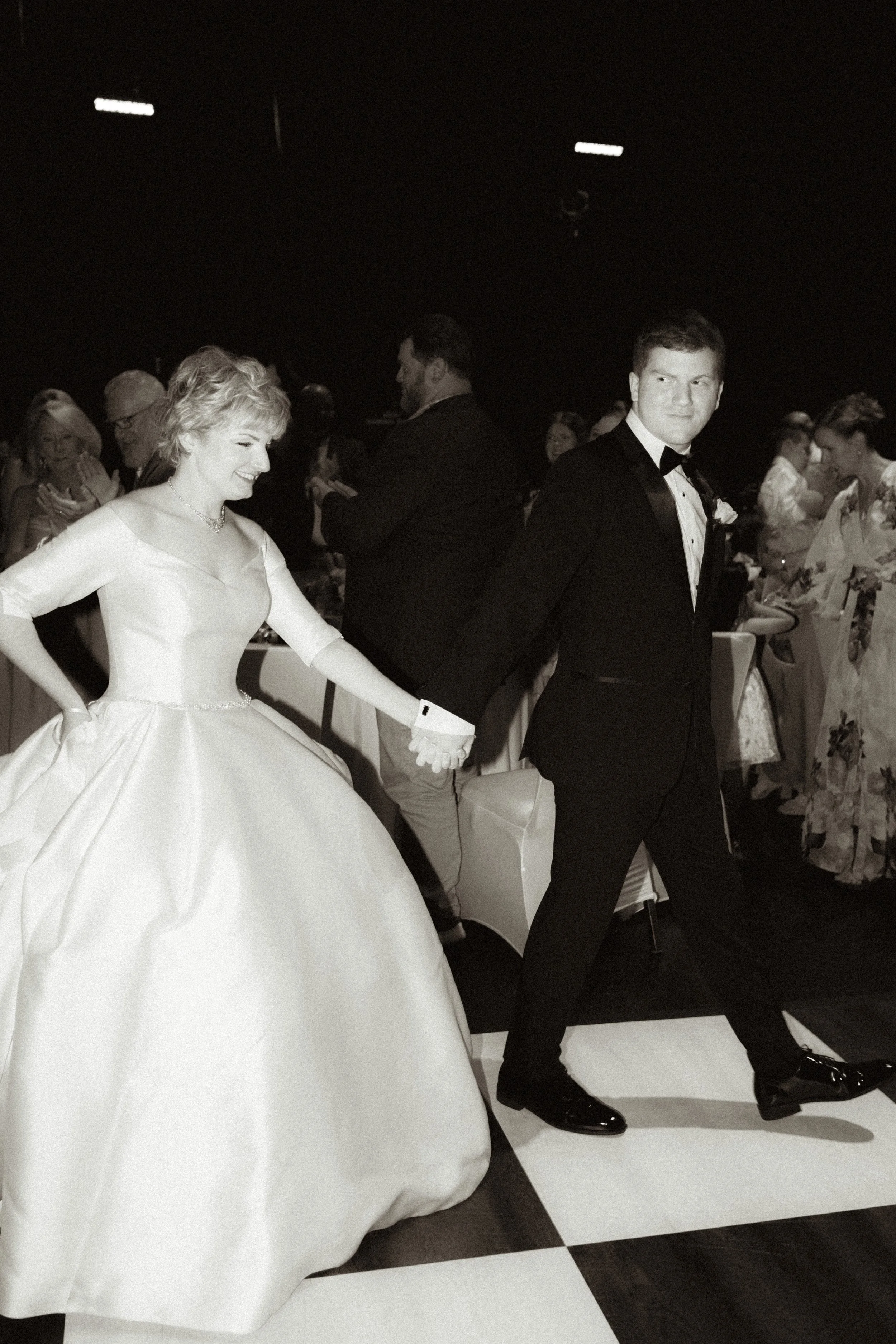 Black and white photo of a bride and groom dancing at their wedding reception, with guests in the background.