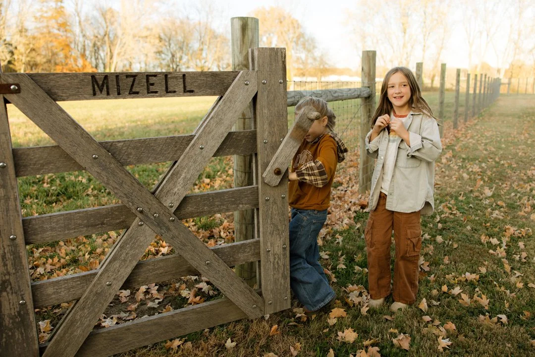 Two children, a boy and a girl, standing outside near a wooden gate with fall leaves on the ground and a fence. The boy is on the other side of the gate, and the girl is smiling while holding her hands near her chest.