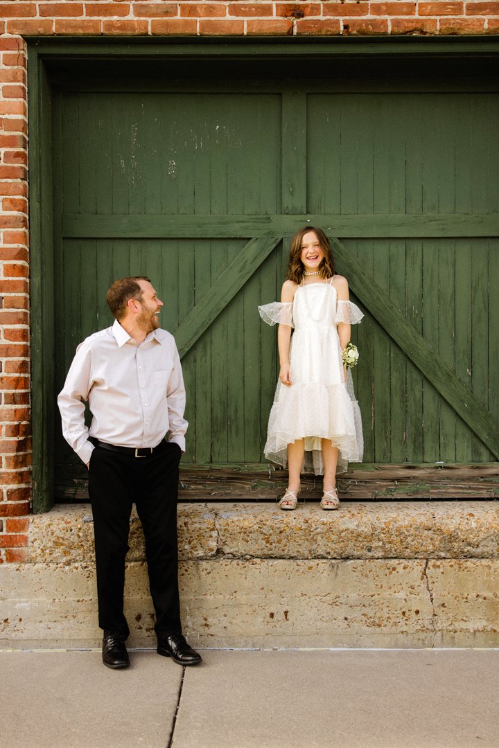 A young girl in a white dress and sandals stands on a wooden platform in front of a large green garage door, smiling and holding a small bouquet. A man in a white shirt and black pants stands nearby, looking at her and smiling. The setting includes a