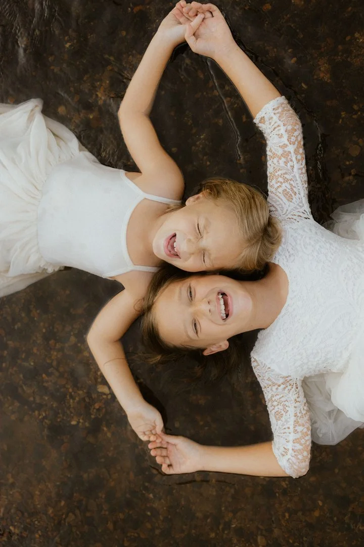 Two young girls lying on a dark marble floor, holding hands, laughing, and wearing white dresses.