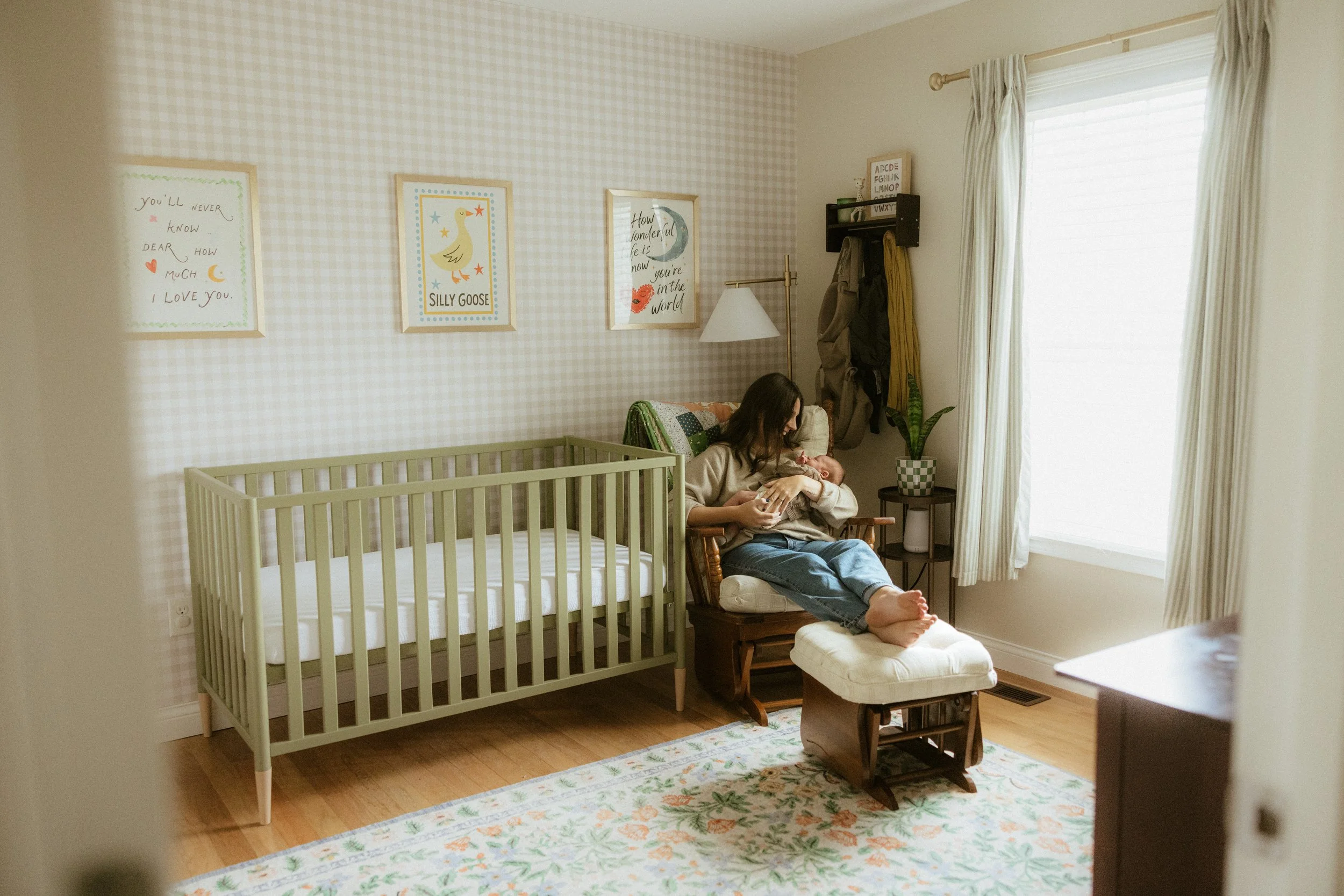 A woman sits in a rocking chair with a baby, in a nursery with a crib, wall art, and a large window with curtains.
