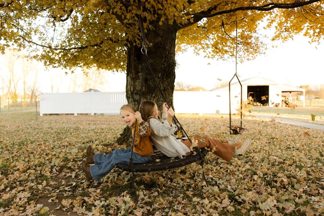 Two children sitting on a large circular rope swing hanging from a tree in a yard during fall. The children are laughing and enjoying the moment as the leaves fall around them.