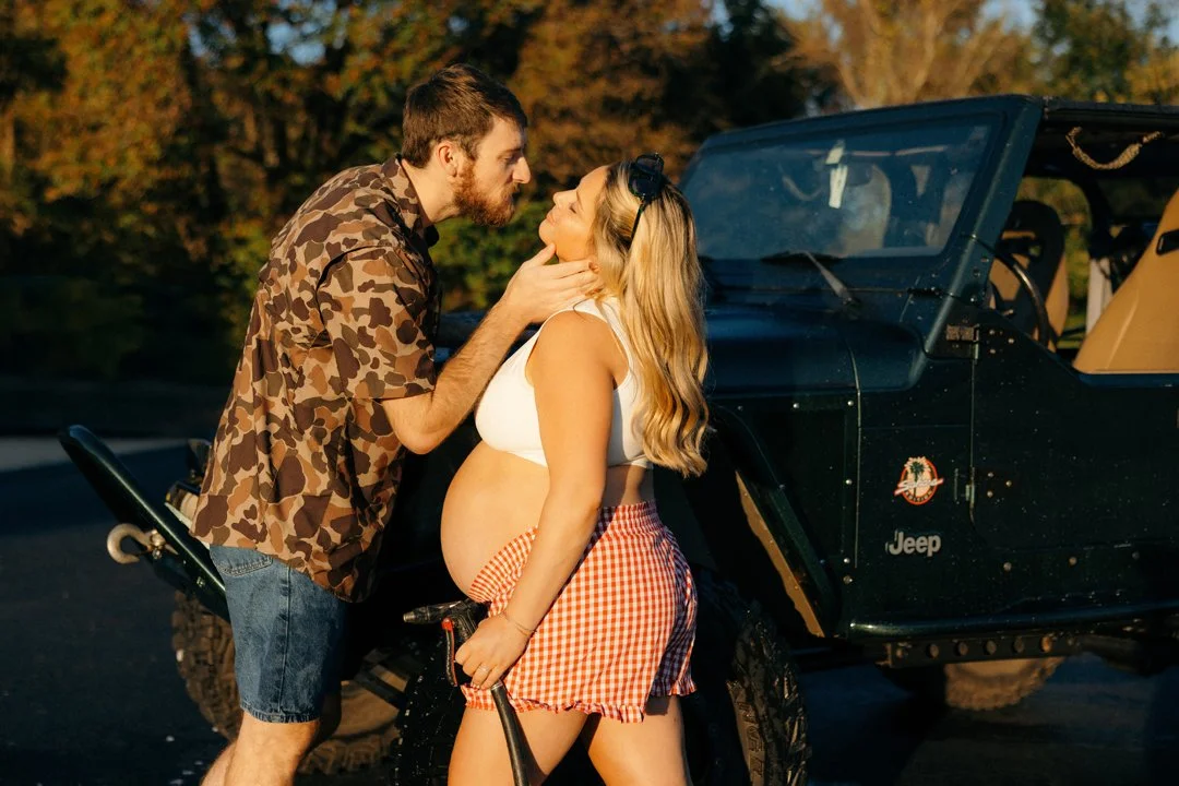 Man and pregnant woman standing close to each other outdoors near a black Jeep, with trees in the background, during sunset.