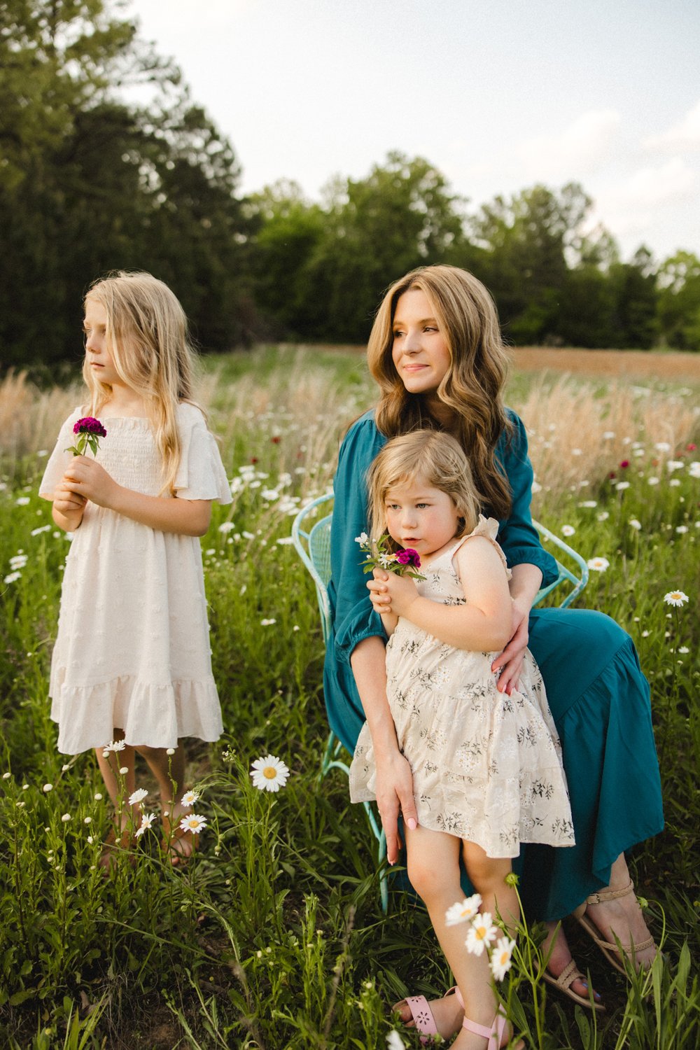 A woman and two young girls in a field of daisies and wildflowers, with trees in the background. The woman is sitting in a blue chair, holding one girl on her lap, while the other girl stands nearby holding a flower.