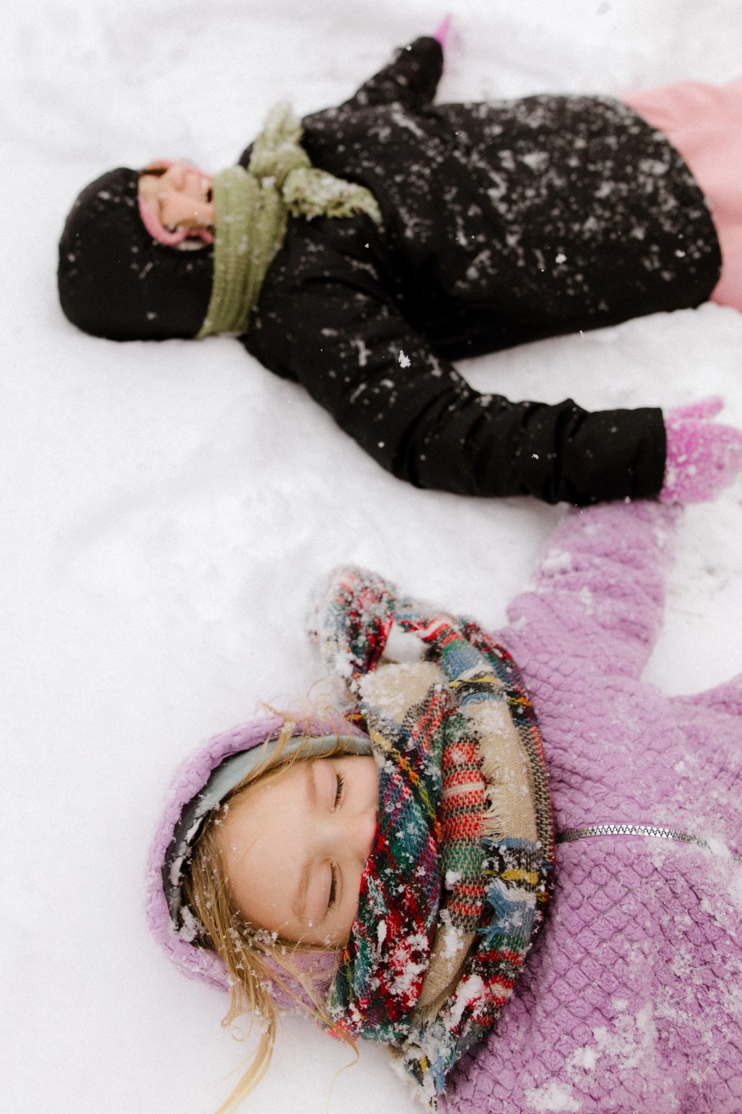 Two children lying on snow, wearing winter clothing, with eyes closed, enjoying snow day.
