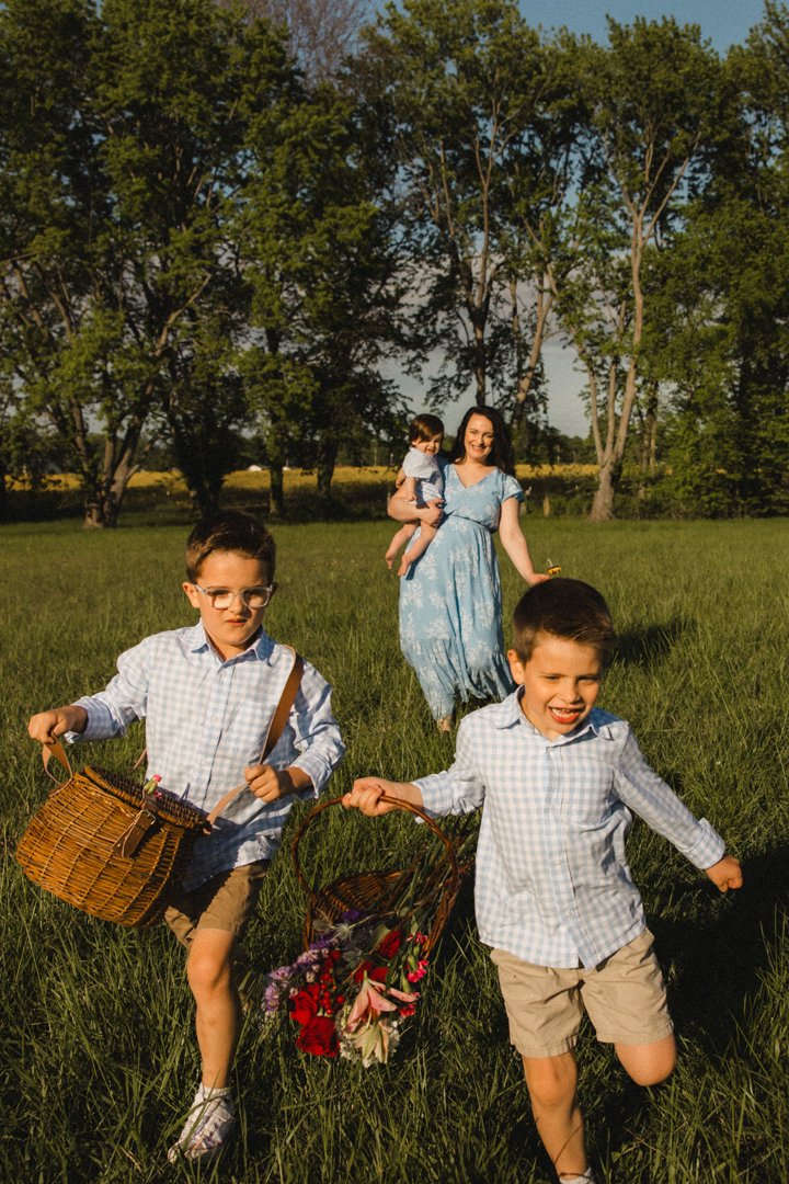A woman and three children enjoying a day outdoors in a grassy field with trees, carrying baskets with flowers, under a clear blue sky.