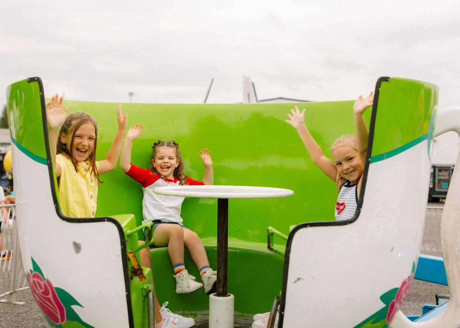 Three young girls smiling and waving inside a colorful kiddie ride shaped like a teacup at an amusement park.