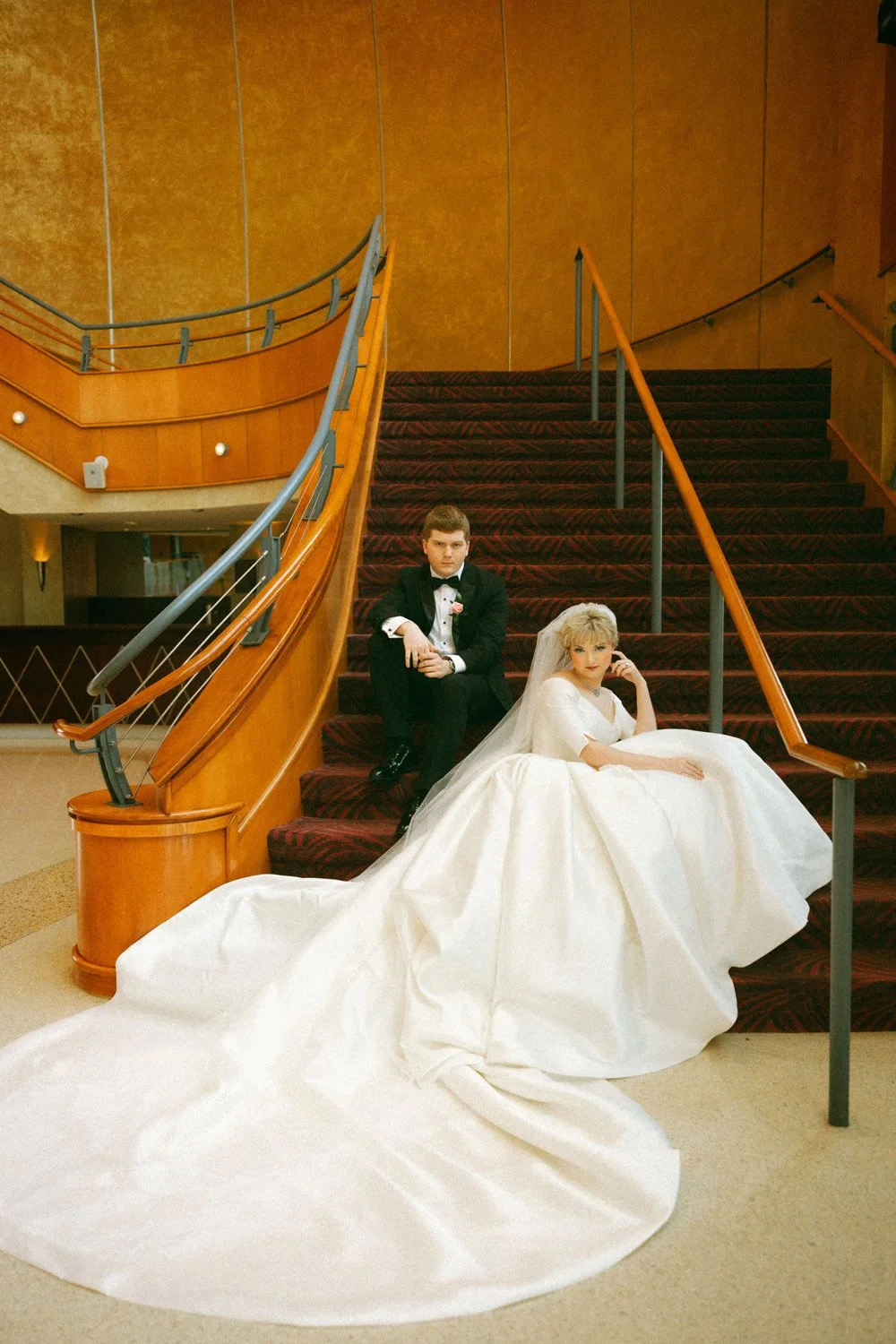 A bride in a white wedding dress with a long train and a groom in a black tuxedo with a bowtie sitting on a staircase with red carpet and wooden railing.