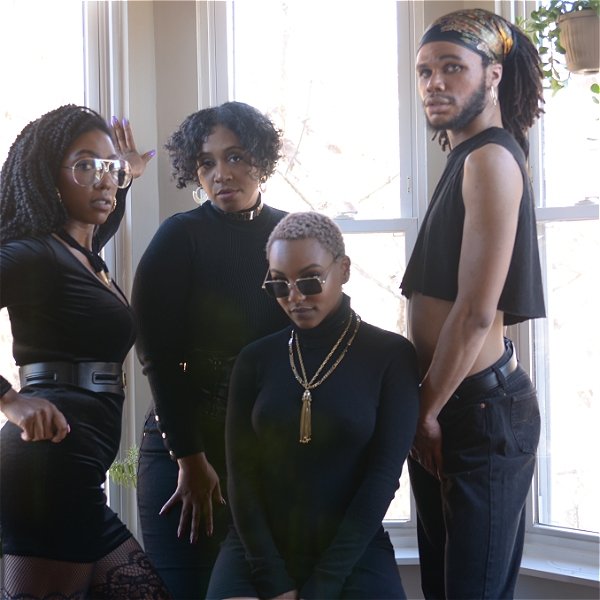 A troupe of four young black adults, dressed in all black, stare back at you, center person in sunglasses