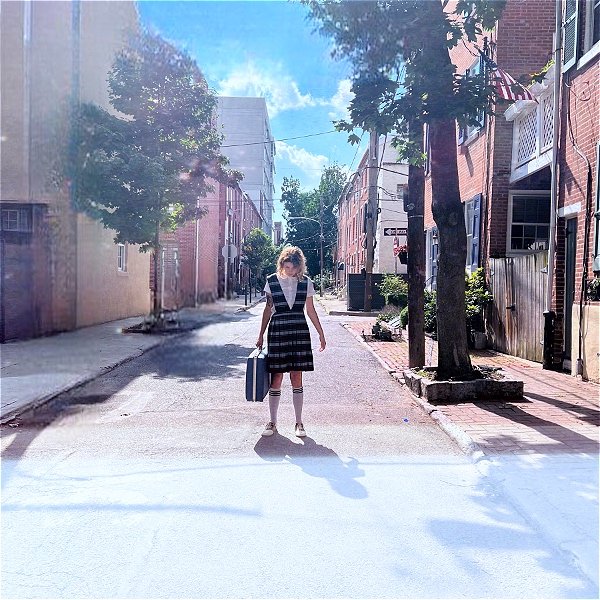 A woman holds a suitcase in the middle of a tree-lined street, looking down