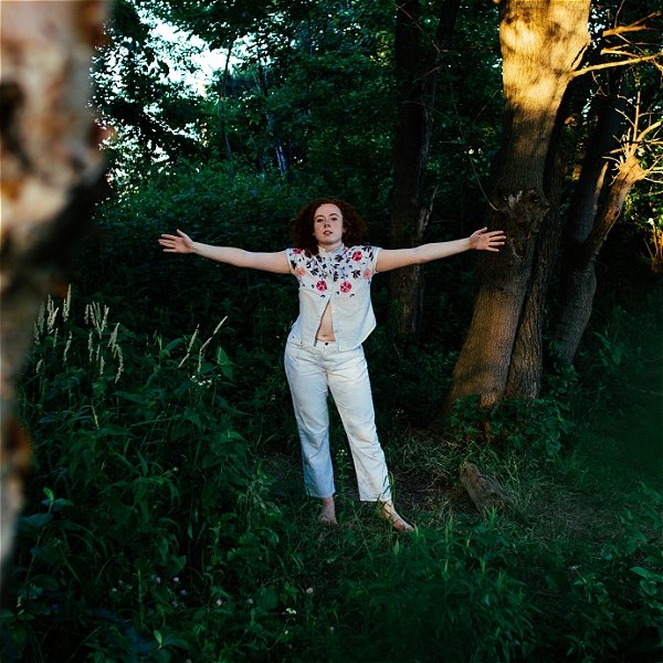 A woman in white stands, arms outstretched, in the woods at magic hour