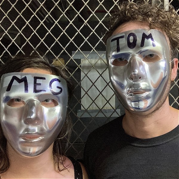 Two actors wear chrome masks labeled MEG and TOM
