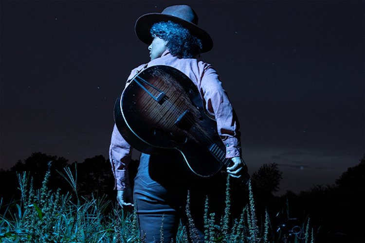 Hatted black cowboy faces away, walking into the grassy plains, towing a guitar on his back
