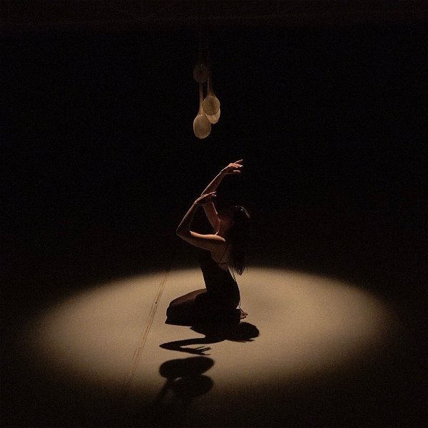 Dancer kneels below suspended dangly frabricky orbs, arms akimbo