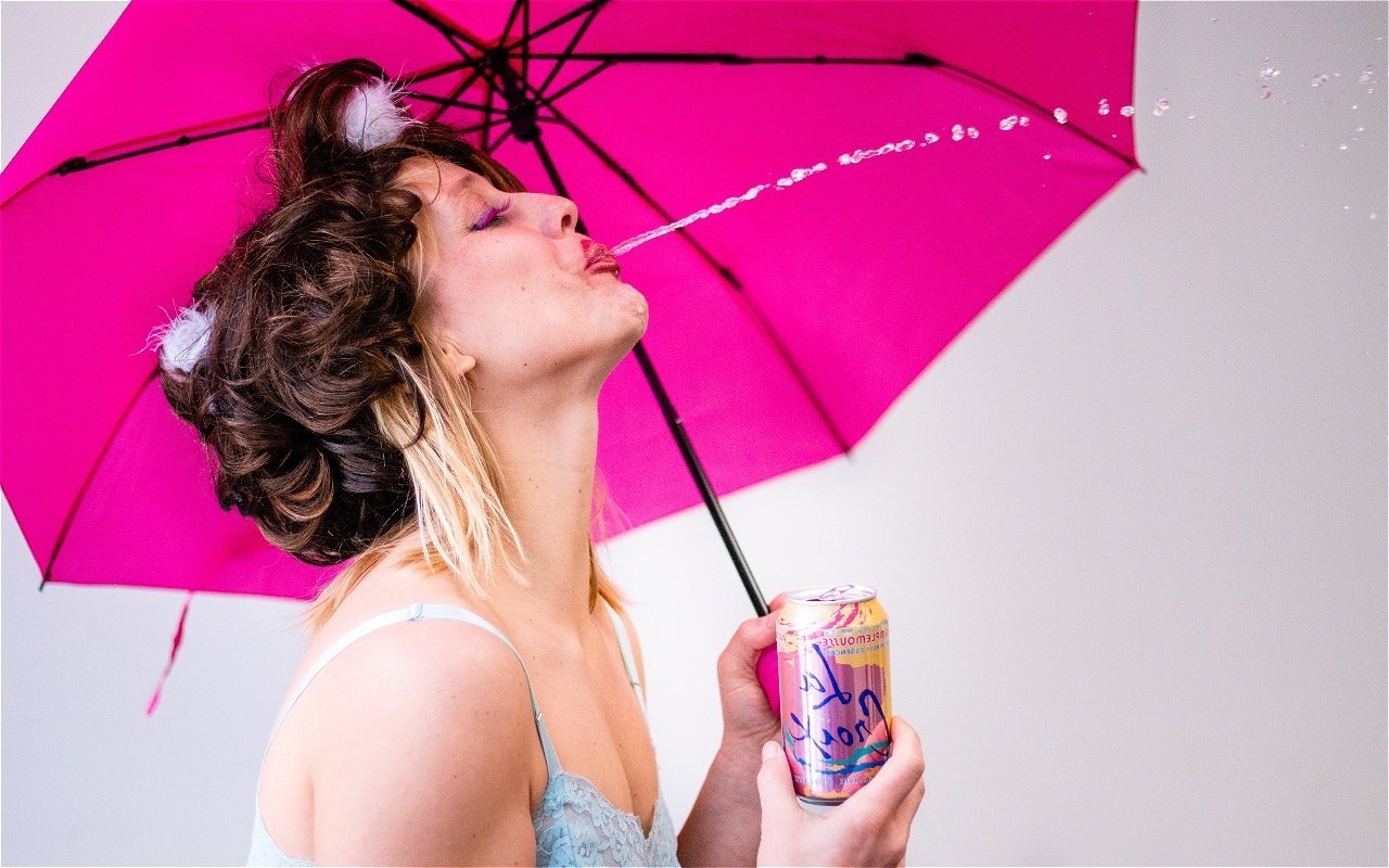 White woman spitting out a stream of La Croix from under a pink umbrella