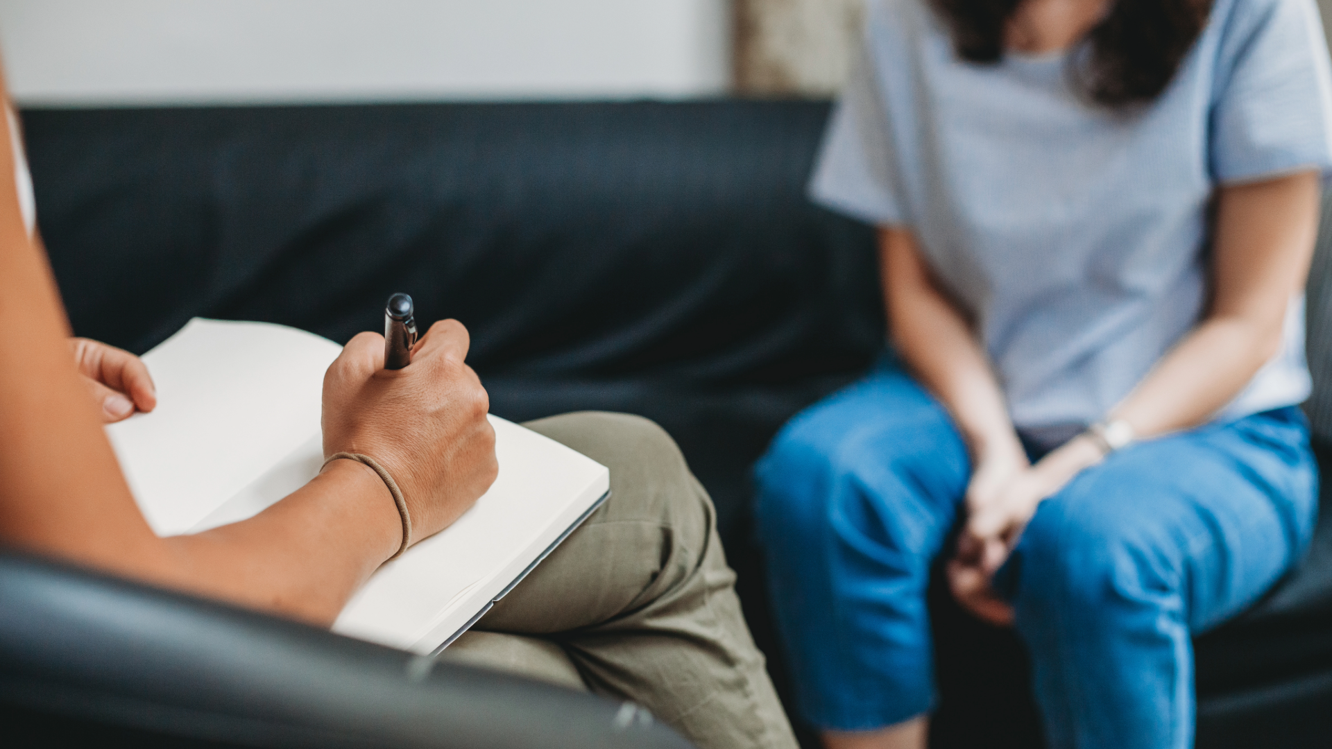 A therapist taking notes with a pen in a notebook during a therapy session or counseling session, with a woman sitting nearby.