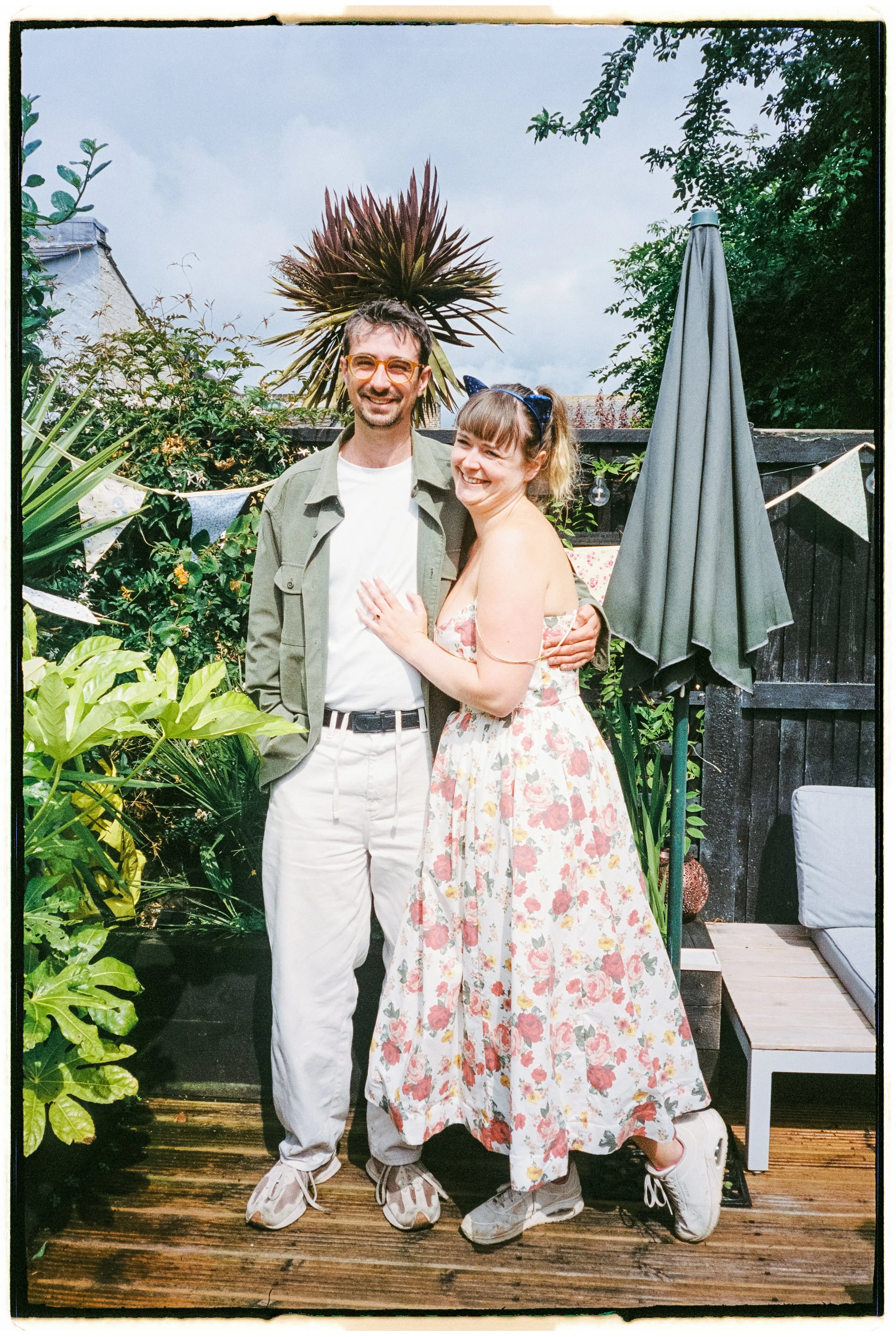 A man and woman smiling and standing close together outdoors in a garden with lush green plants and a black wooden fence. The man is wearing glasses, a light green jacket, white shirt, and white pants. The woman is wearing a floral dress, sneakers, and a navy headband with a bow. There are bunting flags and string lights in the background, along with a large gray patio umbrella.