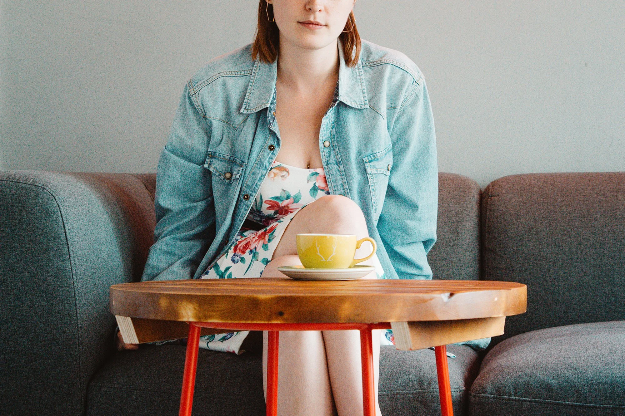 A woman with red hair, wearing a denim jacket over a floral dress, sitting on a gray sofa with her legs crossed. In front of her, there's a round wooden table with a yellow teacup and saucer. The background is a plain light-colored wall.