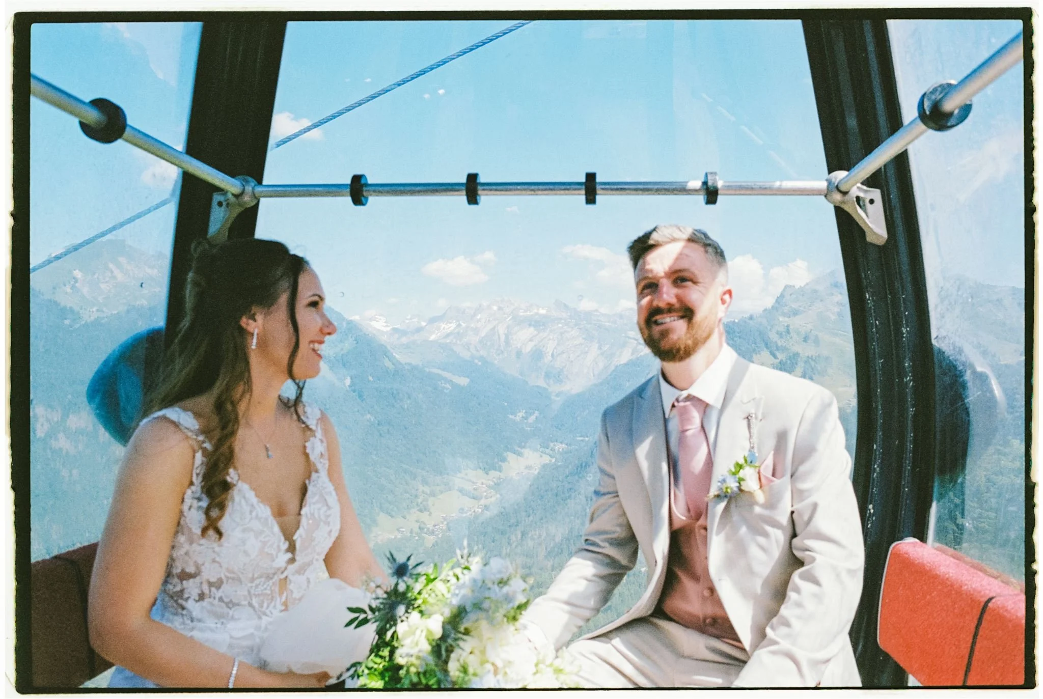 A bride and groom sitting inside a gondola with mountain scenery behind them, dressed in wedding attire. Taken on 35mm film in Morzine, France.