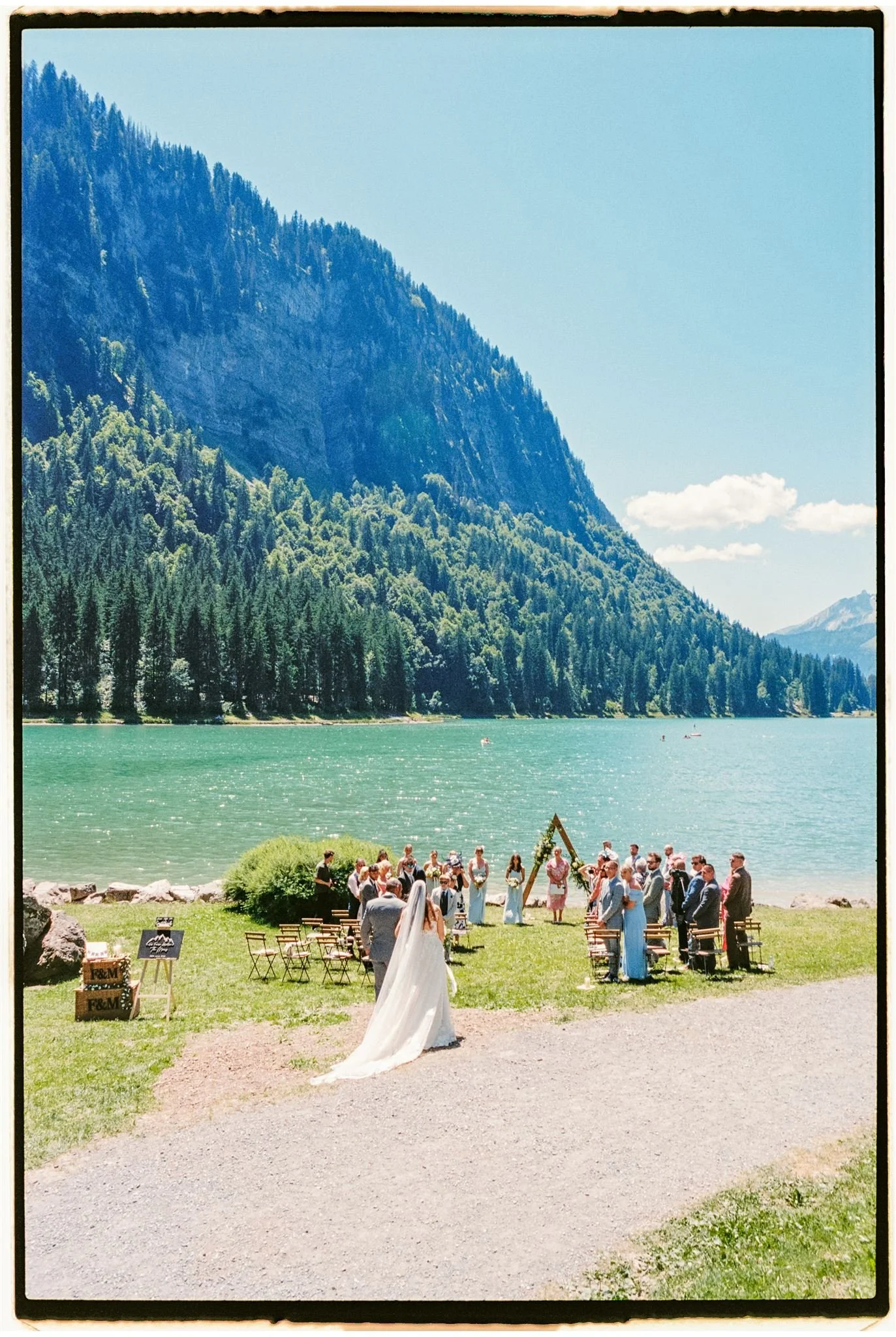 A wedding ceremony taking place outdoors by a lake with a mountain backdrop, with a bride in a white dress and veil, and guests seated on chairs facing an arch. Taken on 35mm film at Lac e Montriond in Morzine, France