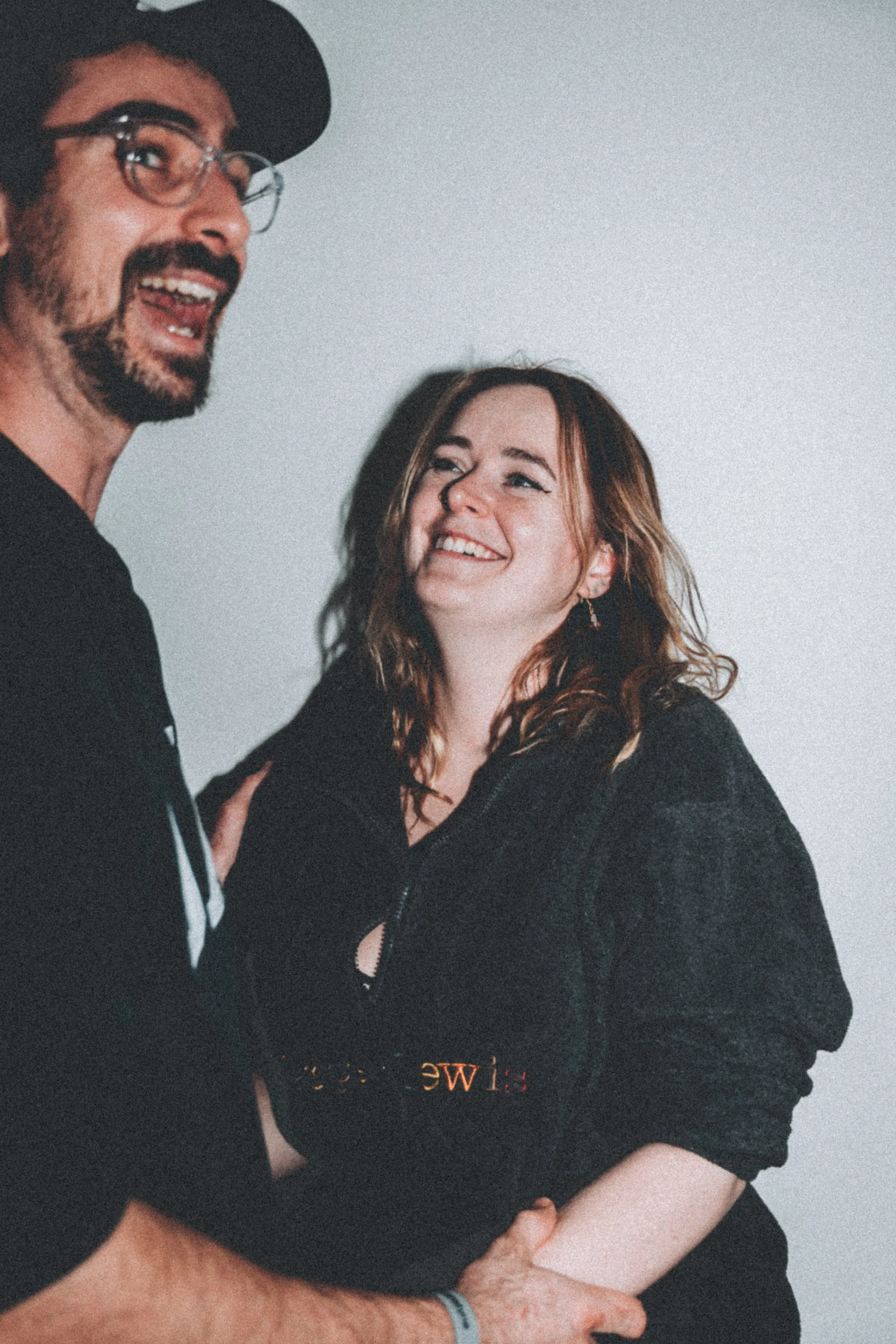 A man with glasses and a beard smiling and a woman with red hair smiling, indoors against a plain light-colored wall.