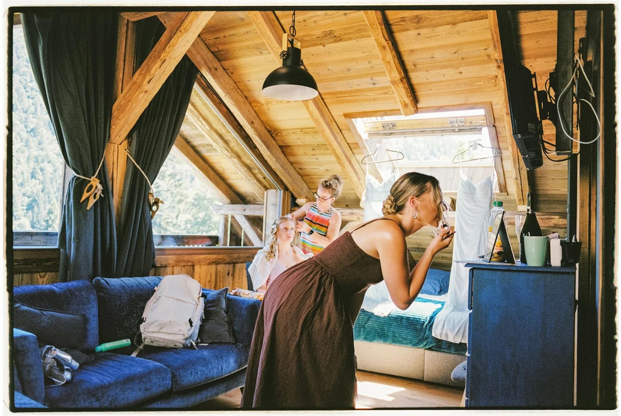 Three women in a rustic, wooden attic room, one applying makeup at a mirror, another sitting in a chair, and the third standing behind her. There are wedding dresses hanging near the window, with natural light streaming in.
