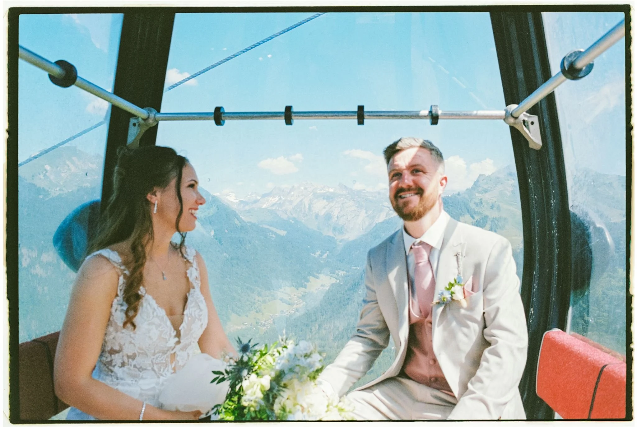 A bride and groom smiling and holding hands inside a cable car with mountain scenery in the background.