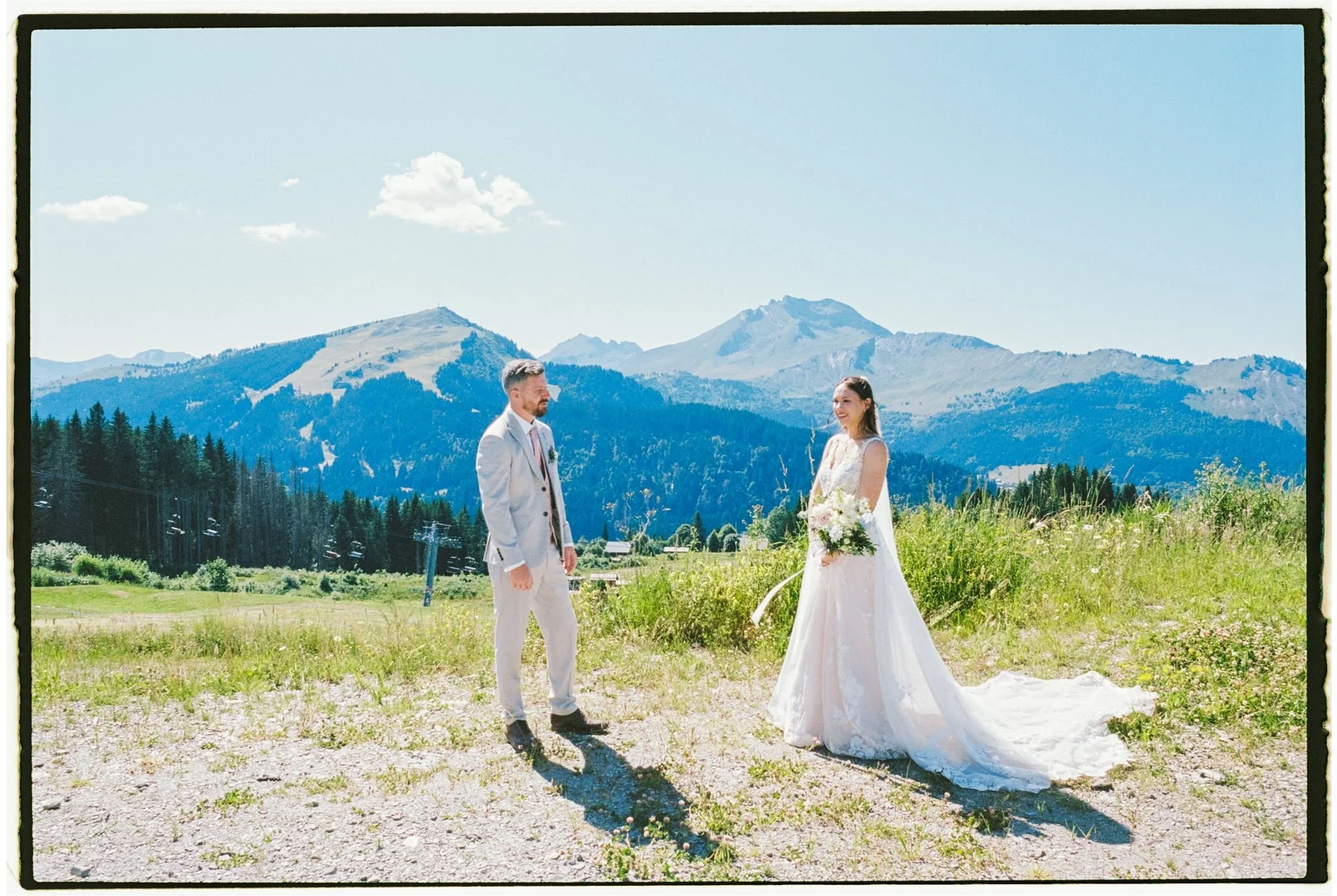A bride and groom standing outdoors on a sunny day with mountains and trees in the background. The bride wears a white wedding gown and holds a bouquet, while the groom wears a light-colored suit.