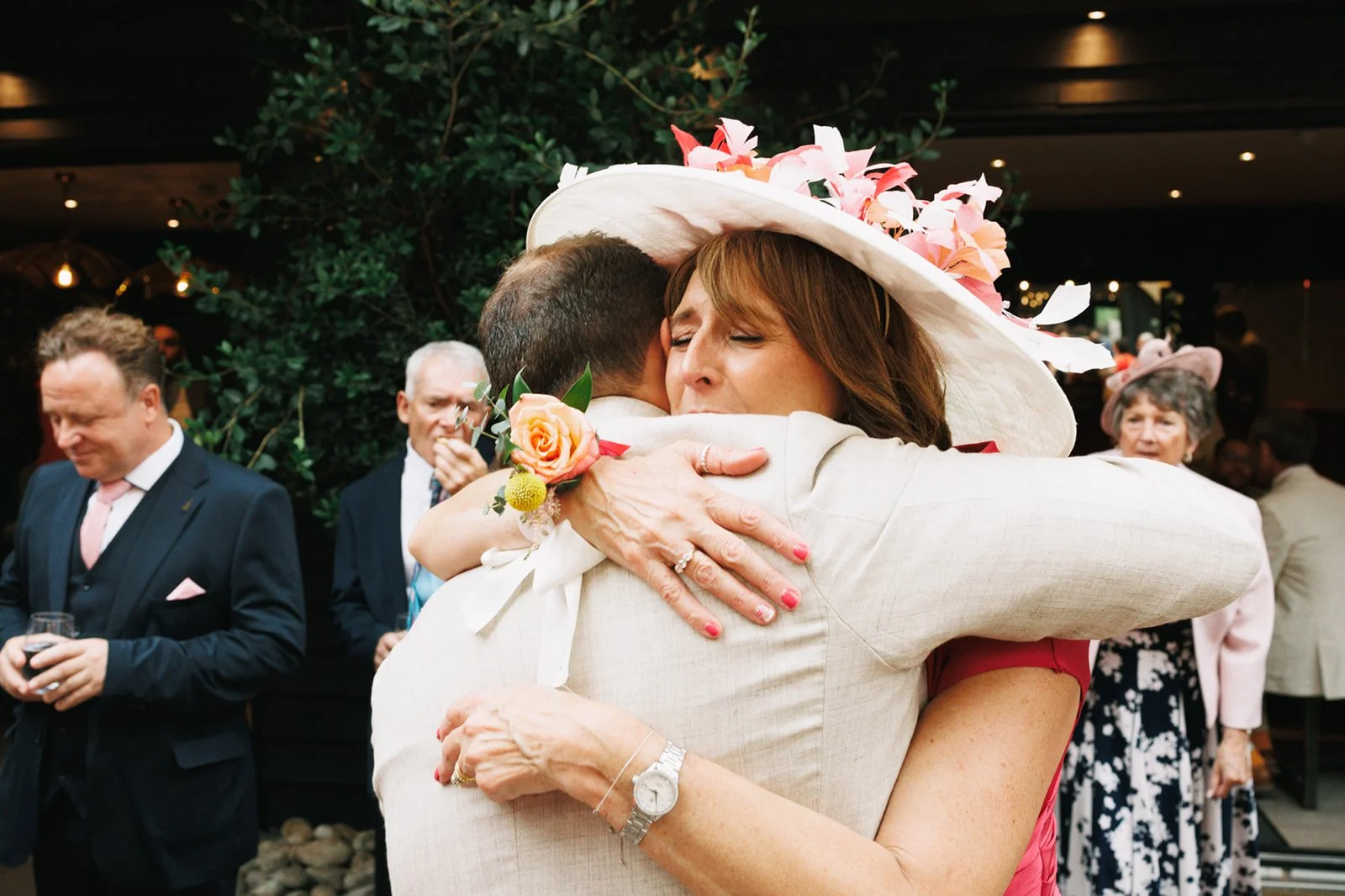 A woman with a floral hat and a woman with painted nails hug at a wedding reception, surrounded by well-dressed guests.
