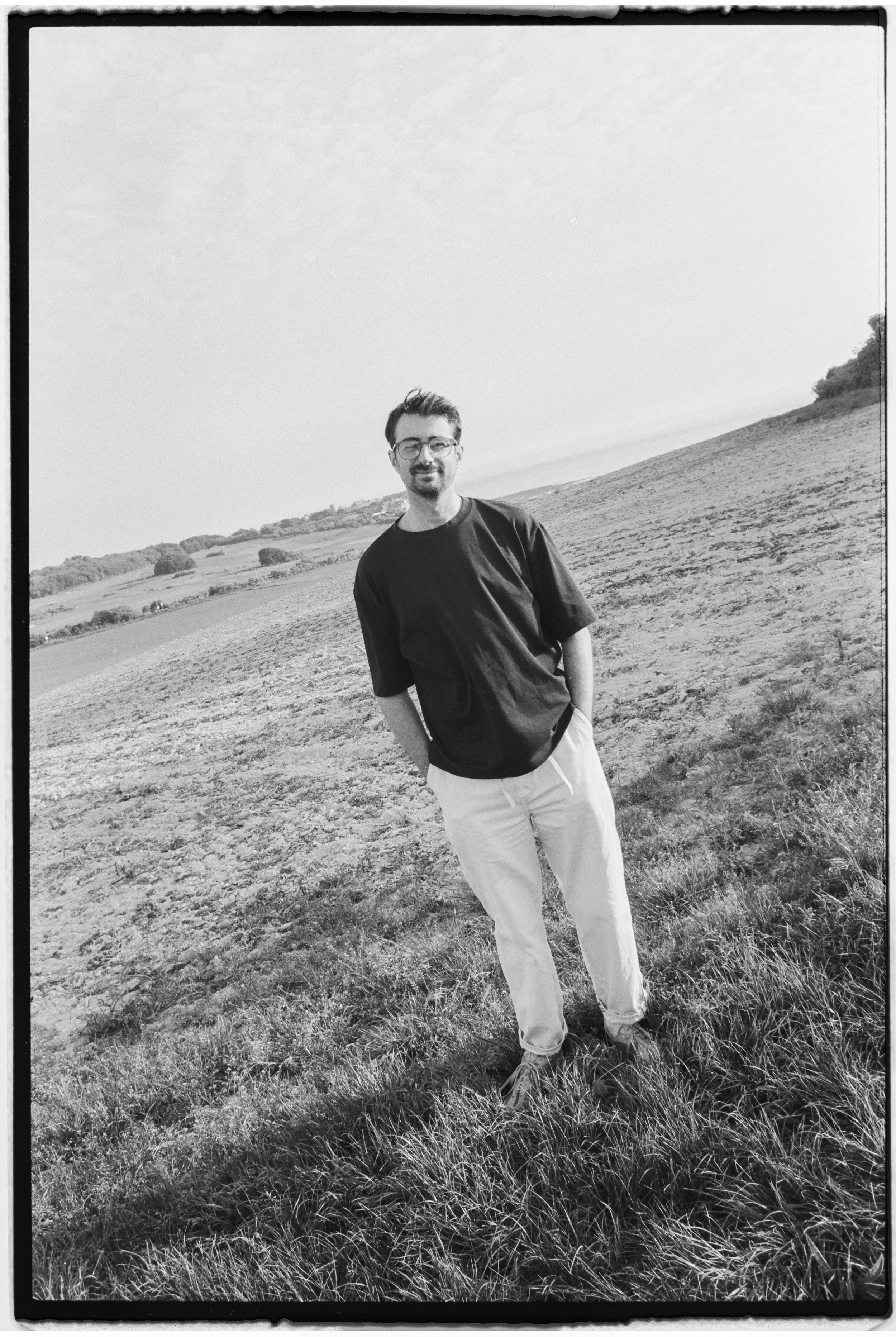A man with glasses and a beard standing outdoors on a grassy hill, smiling at the camera, in black and white.