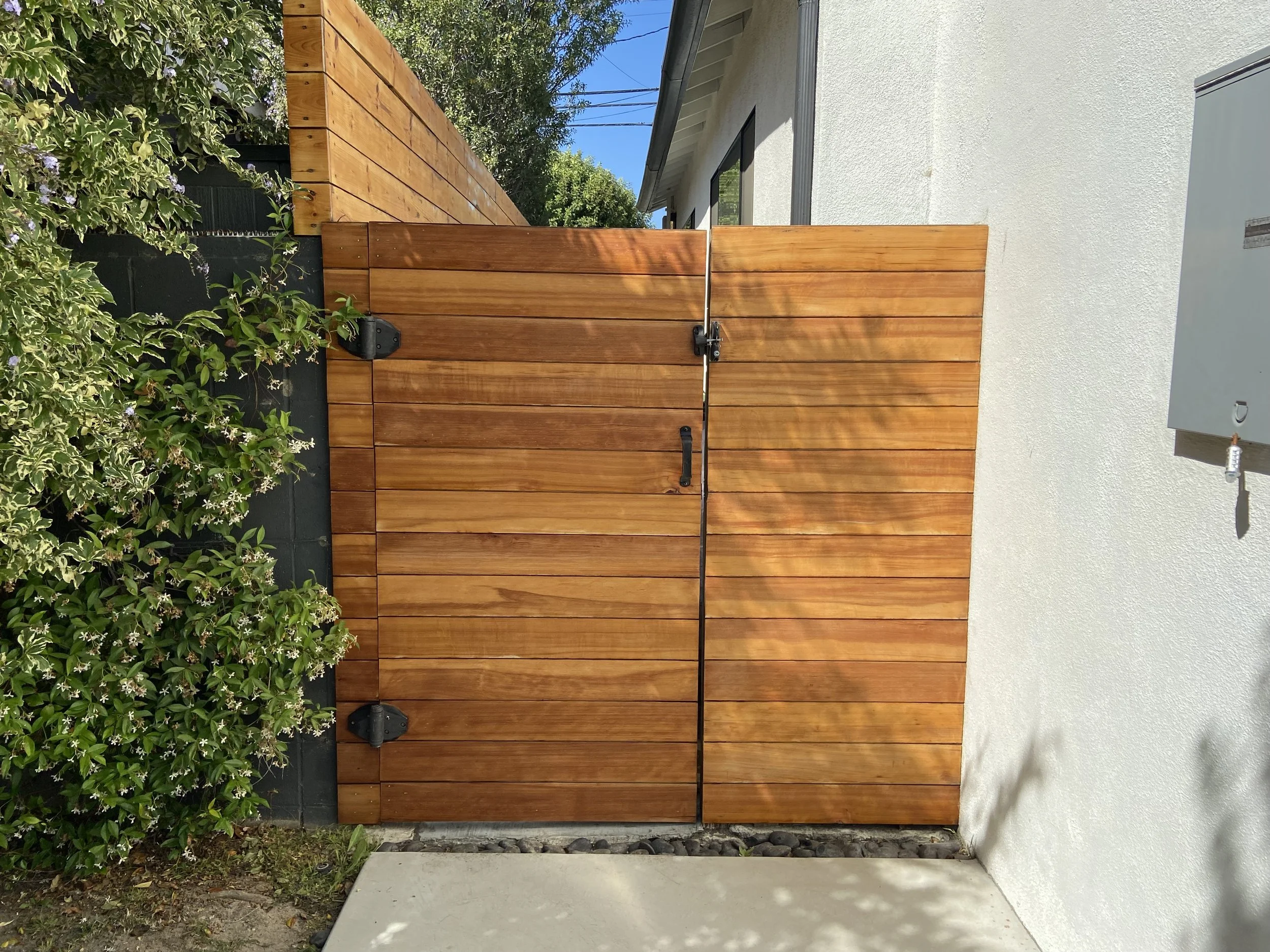 Horizontal wood-plank garden gate with black hardware beside a white stucco house and flowering greenery.
