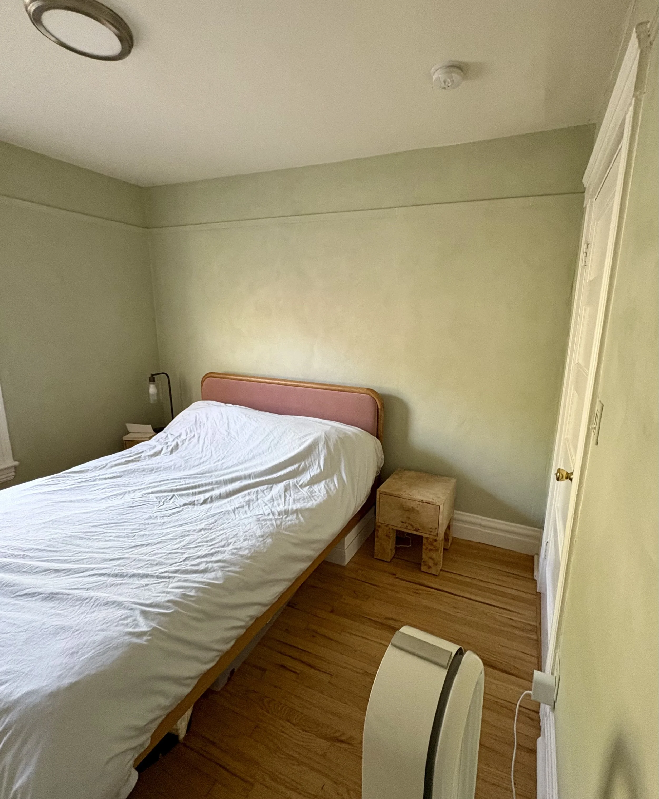 Small bedroom with a white-covered bed, pink upholstered headboard, wooden nightstand, hardwood floors, and an open door.
