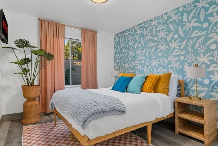 Bright modern bedroom with blue floral accent wall, white bed, colorful pillows, peach curtains, woven nightstand, and potted monstera plant