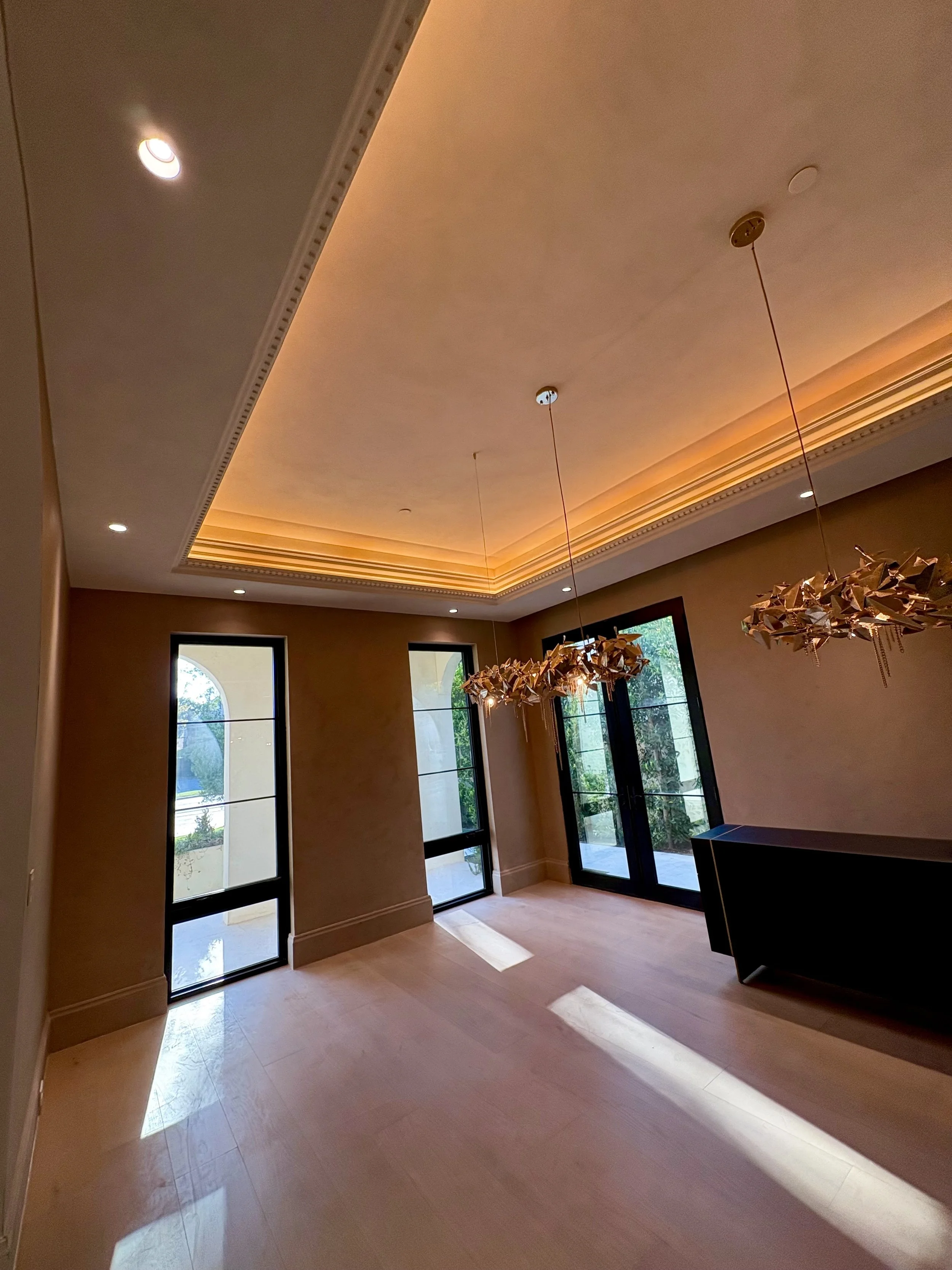 Elegant interior room with a coffered ceiling, warm recessed lighting, tall black-framed windows, and a decorative branch chandelier.