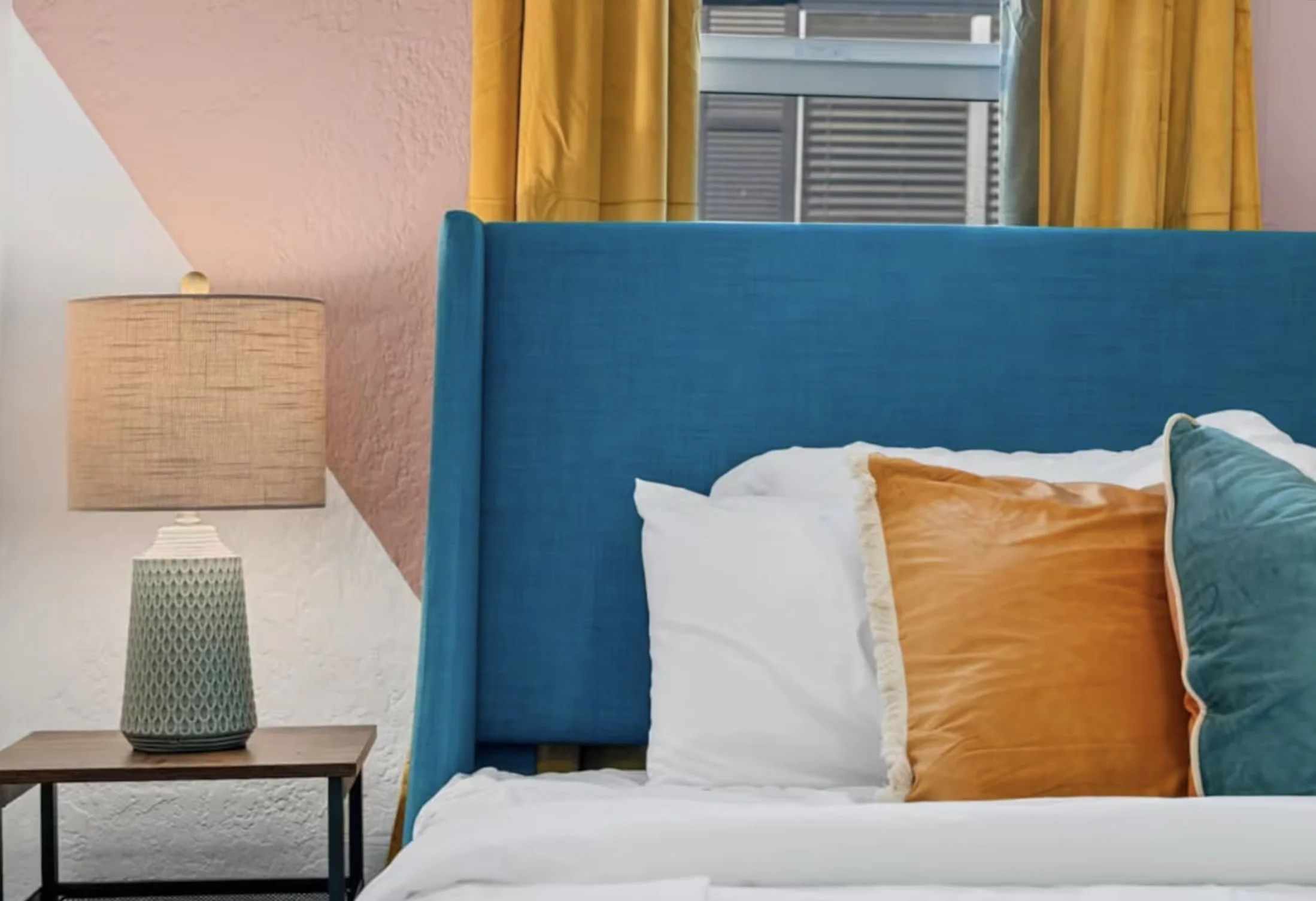 Colorful bedroom with a teal bed, white bedding, orange and teal pillows, mustard curtains, and a bedside lamp.
