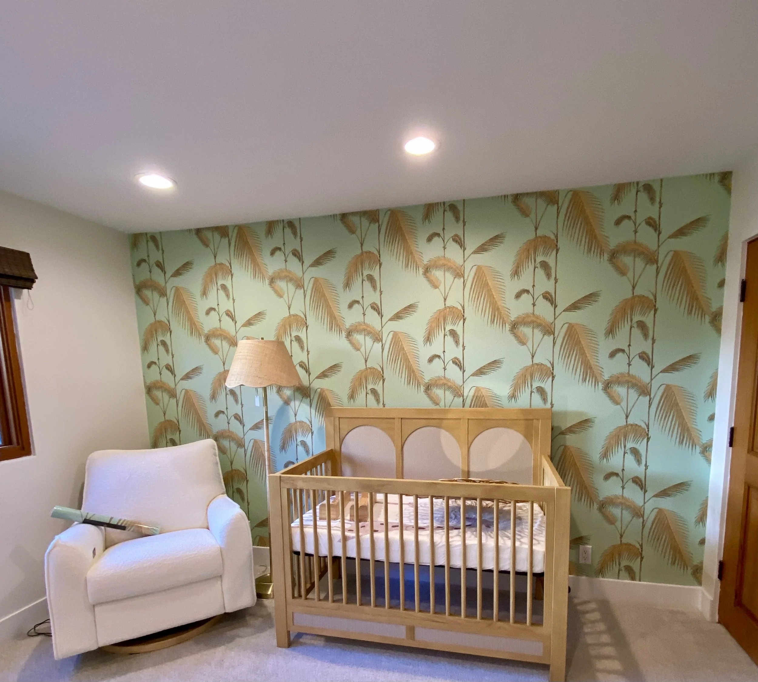 Nursery with a wooden crib, white rocking chair, floor lamp, and tropical palm tree wallpaper