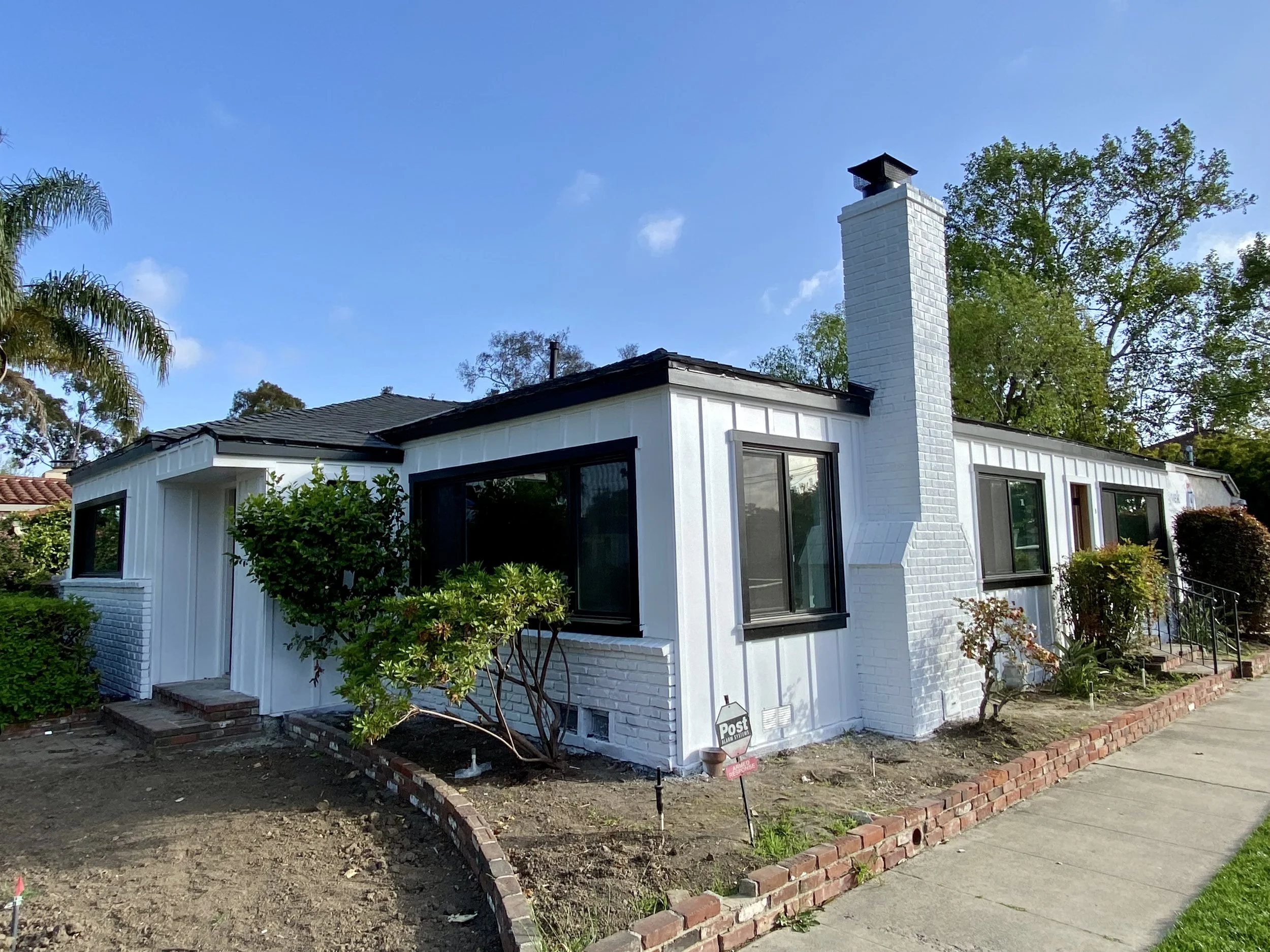 White brick single-story house with black-trimmed windows, chimney, landscaped yard, and security signs under a blue sky