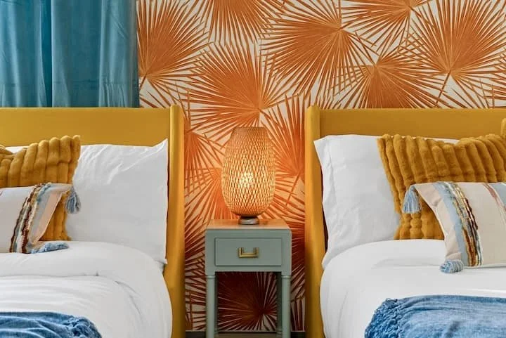 Colorful bedroom with twin beds, mustard yellow headboards, orange palm-leaf wallpaper, glowing bedside lamp, and blue accents
