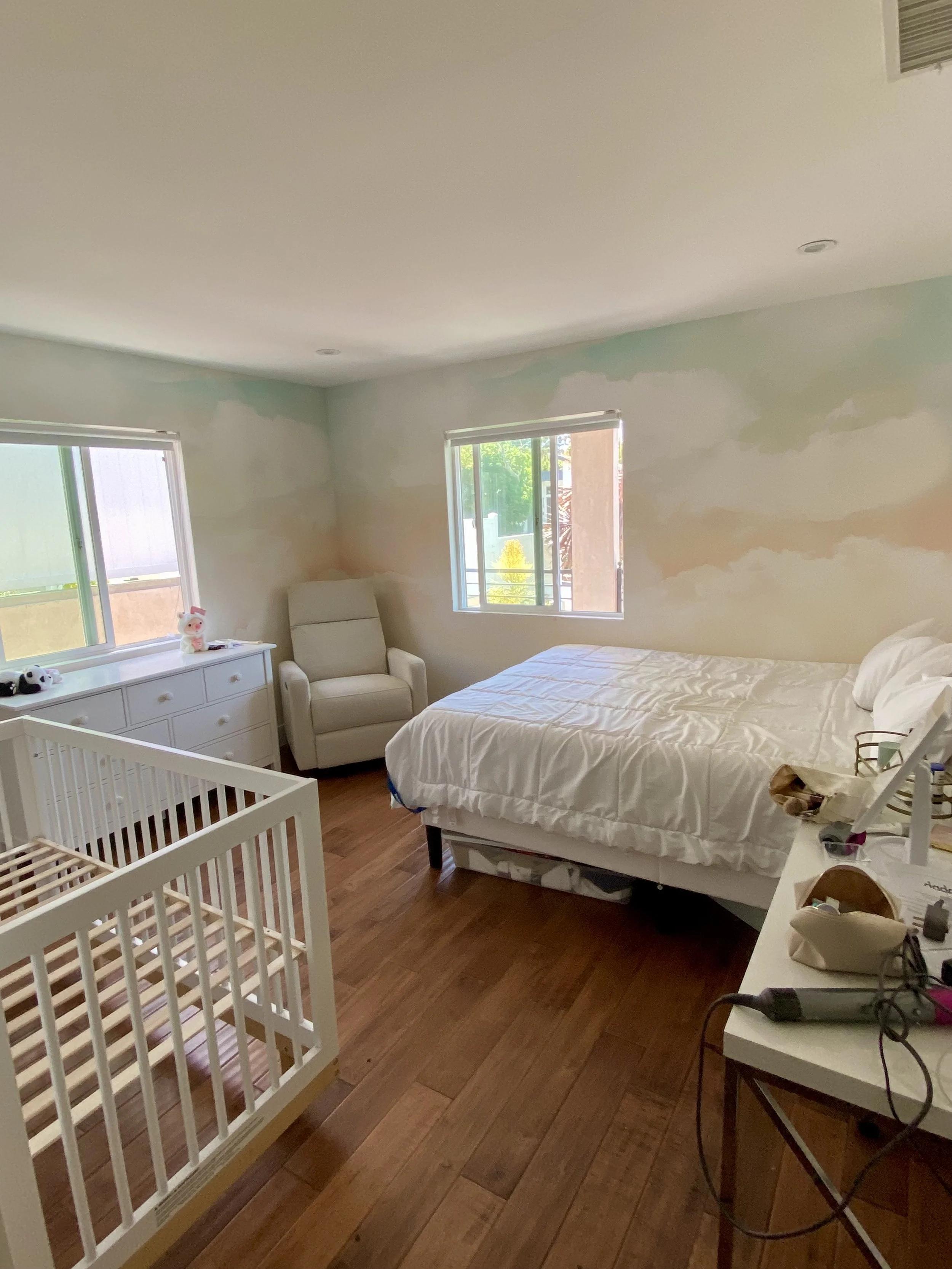 Bright bedroom with a white bed, baby crib, dresser, armchair, windows, and hardwood floors.