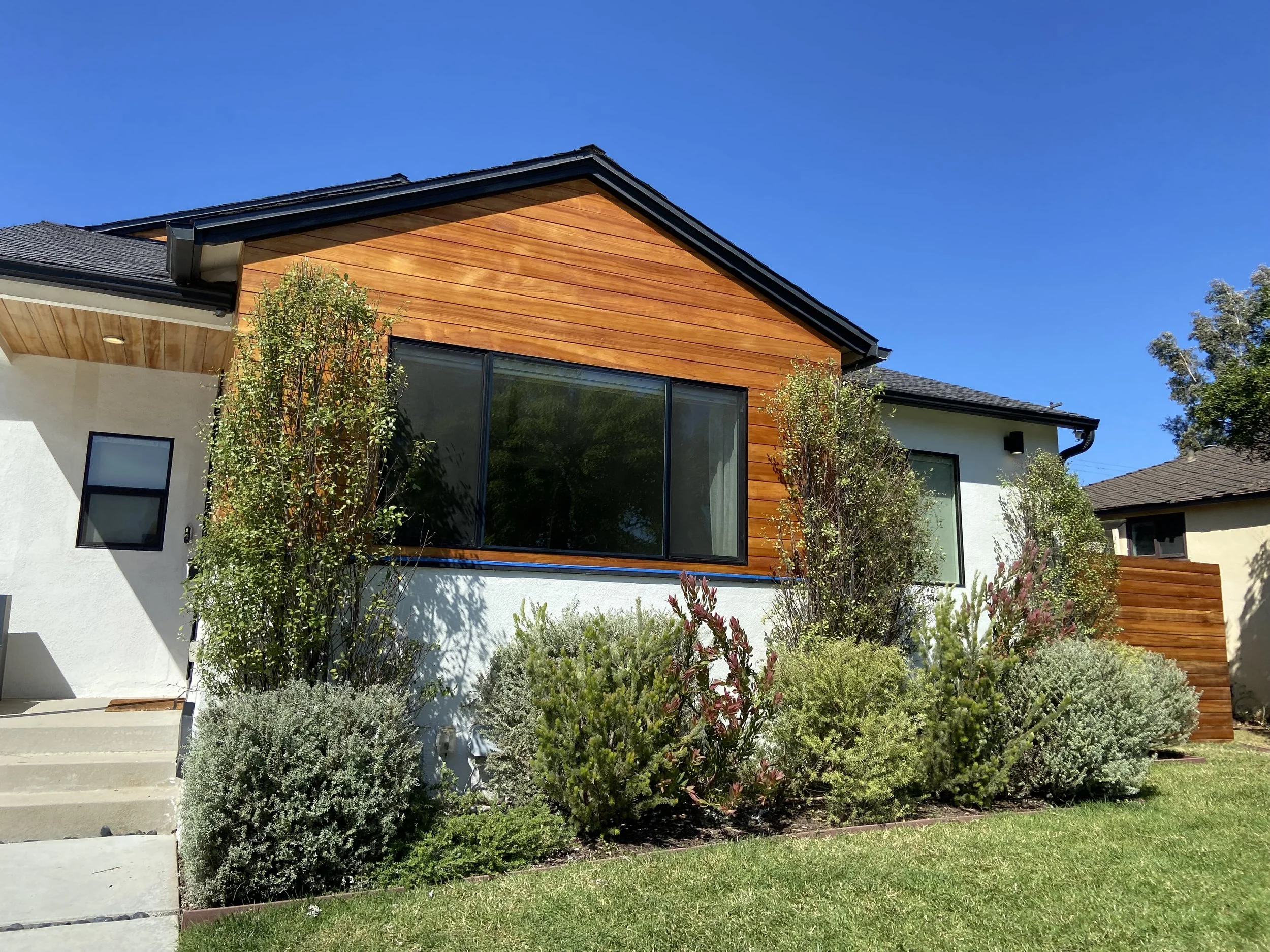 Modern single-story home with cedar siding, large black-framed windows, landscaped front yard, and blue sky