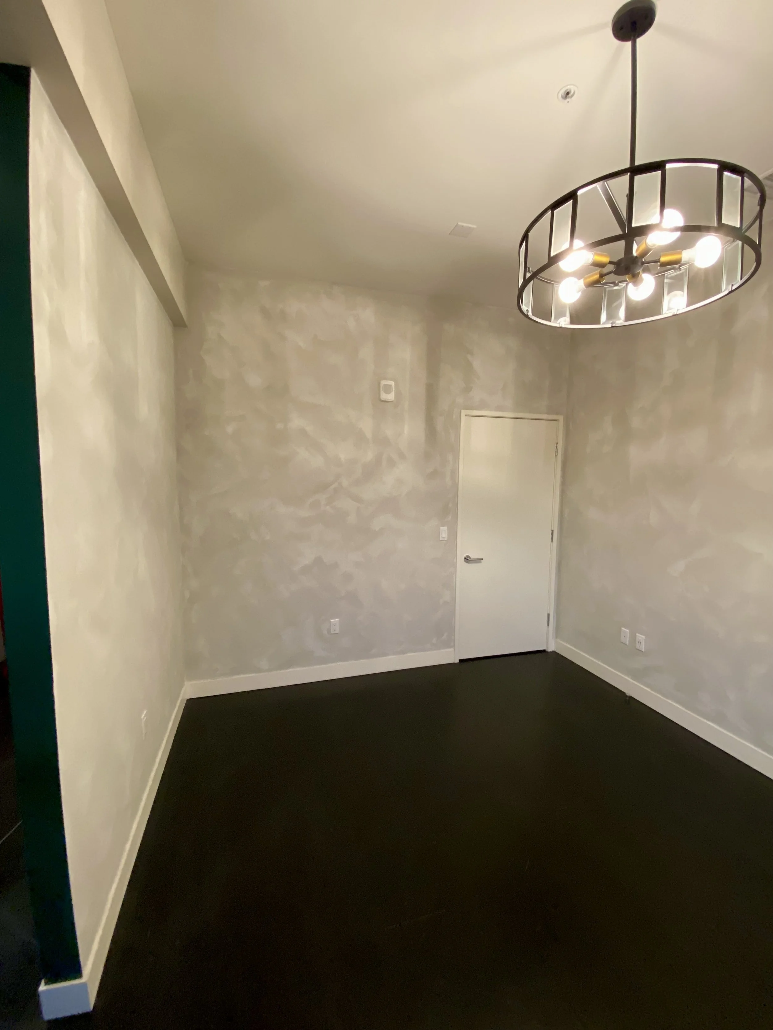 Empty room with unfinished textured walls, dark carpet, a white door, and a modern black-and-brass chandelier.